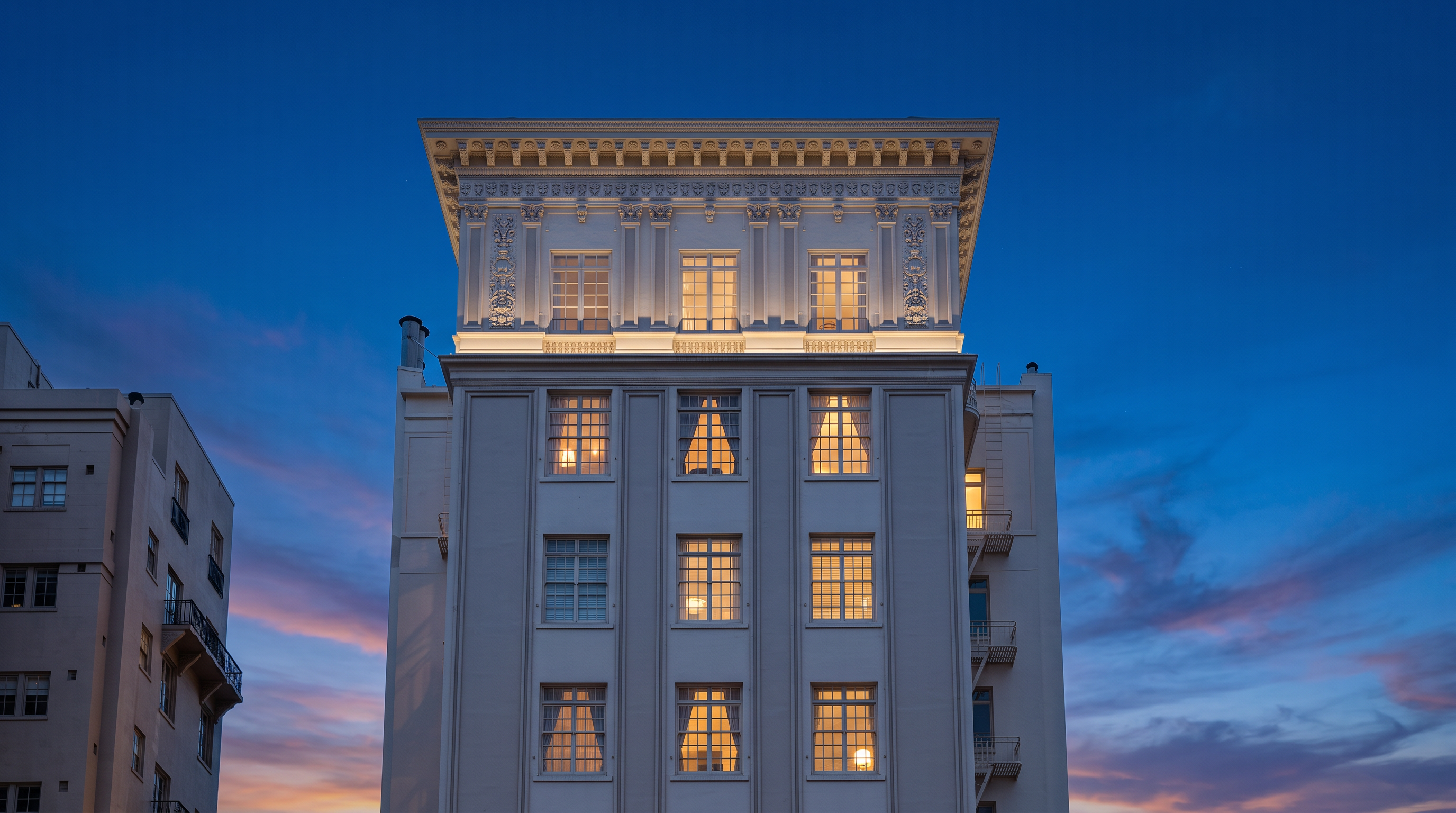 This image showcases the front facade of a tall, historic-style urban building featuring symmetric vertical columns and grid-patterned windows. The architectural design includes an ornate, classic cornice at the top, contributing to a sense of sophisticated, traditional urban living. Its prominent, light-colored facade stands out clearly against the bright blue sky.
