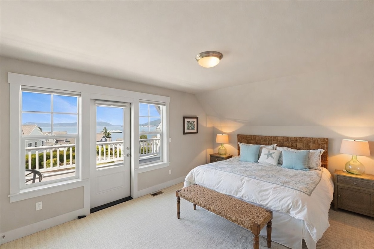 This bright and airy primary bedroom features a king-sized bed with a woven headboard and a coastal-inspired bedding set. A glass door flanked by two large windows provides direct access to a balcony with expansive water views, filling the room with natural light. The space is finished with neutral carpeting, a wooden bench at the foot of the bed, and matching bedside lamps, creating a serene and inviting atmosphere.