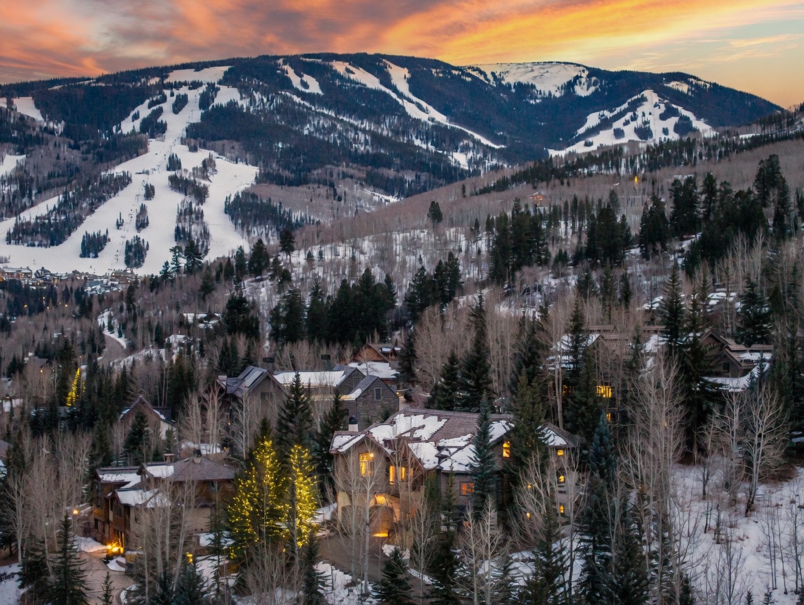 This elevated aerial view captures a luxurious mountain residence nestled within a dense, snow-dusted forest at twilight. The home features a multi-gabled roofline and warm, inviting interior lighting that contrasts beautifully with the dramatic, fiery sunset sky and the expansive, snow-covered ski slopes in the background. The scene conveys a sense of serene, high-end mountain living, emphasizing the property's prime location and architectural integration with the rugged landscape.