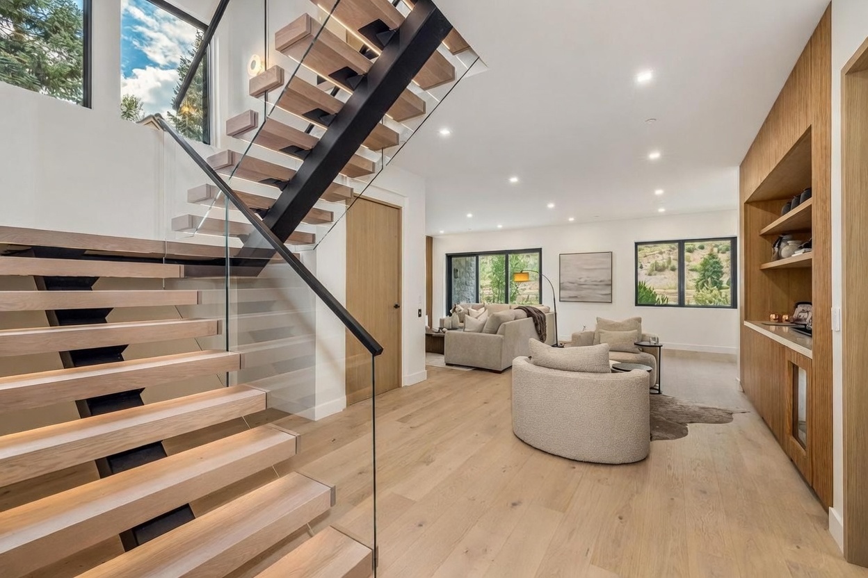 This bright, open-concept interior features a striking floating staircase with light wood treads and a sleek black metal frame, complemented by a glass railing. The perspective captures the transition from the entryway into a cozy living area, which is furnished with neutral-toned seating and accented by a built-in light wood shelving unit along the wall. Large windows provide ample natural light and views of the surrounding landscape, creating an airy and modern atmosphere.