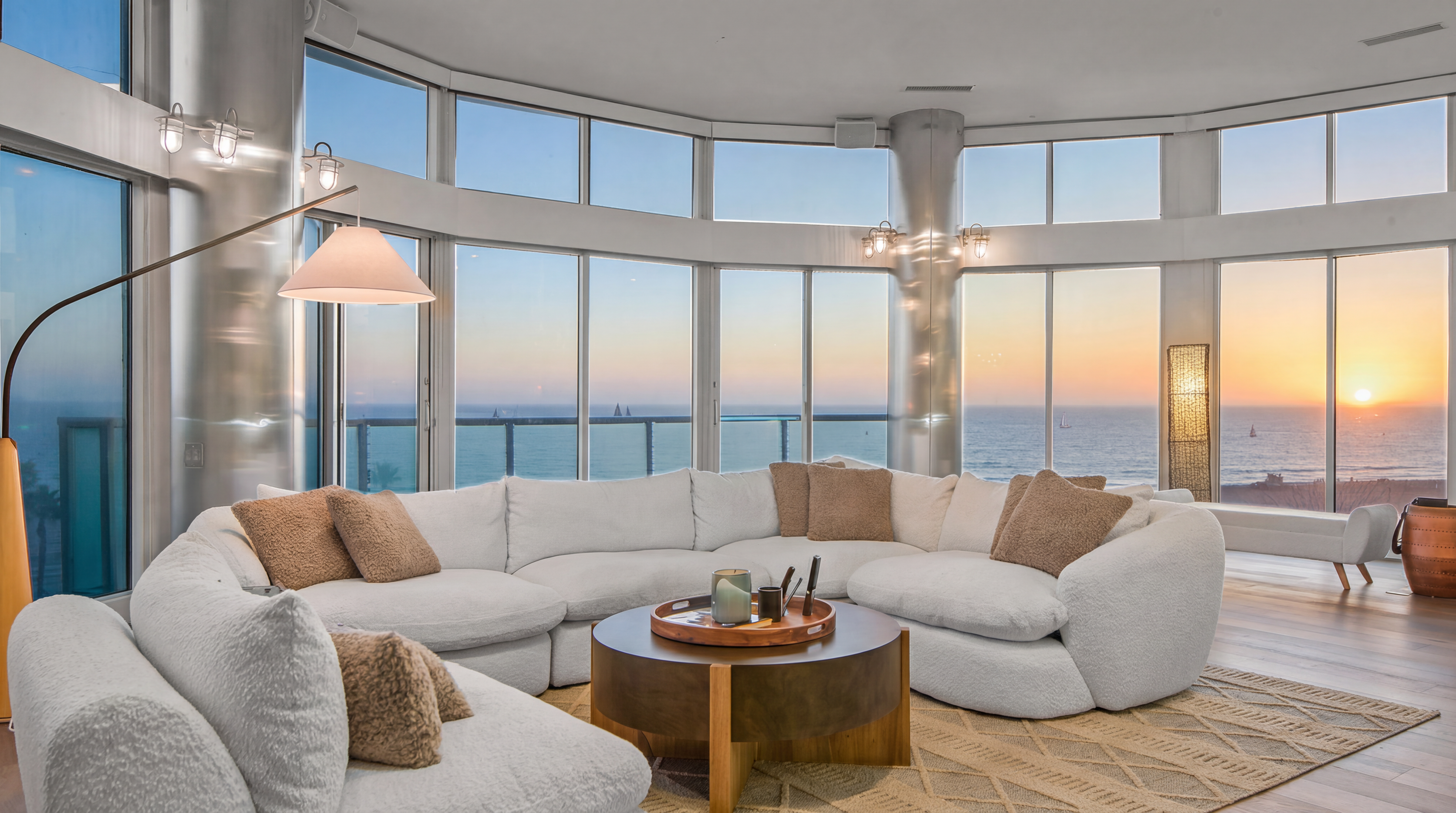 This modern living room features a large, plush white sectional sofa centered around a circular low-profile coffee table on a textured area rug. The space is defined by its expansive, curved floor-to-ceiling windows that provide panoramic sunset views of the ocean. High-end architectural elements like polished metallic structural pillars and sleek recessed lighting complement the sophisticated and airy atmosphere.