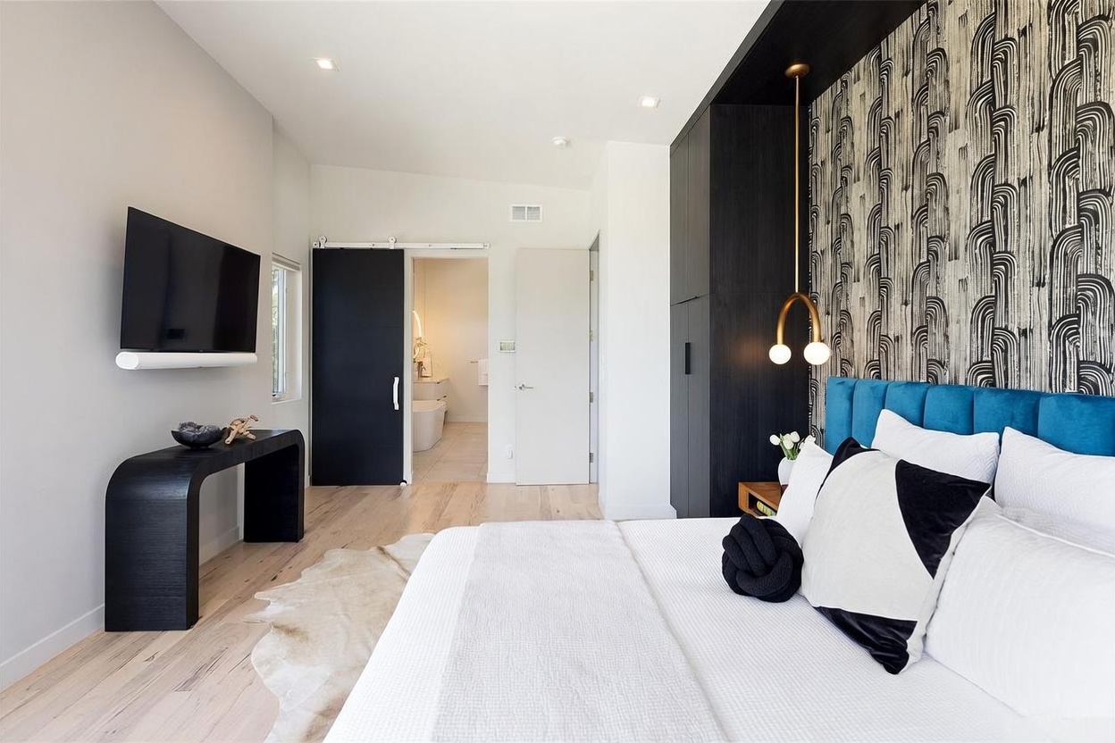This modern primary bedroom features a bold accent wall with patterned wallpaper behind a plush blue upholstered headboard. A sleek black console table sits beneath a wall-mounted television, while a sliding black barn door provides access to an adjacent bathroom. The room is finished with light wood flooring, a textured area rug, and a unique gold-toned pendant light fixture.