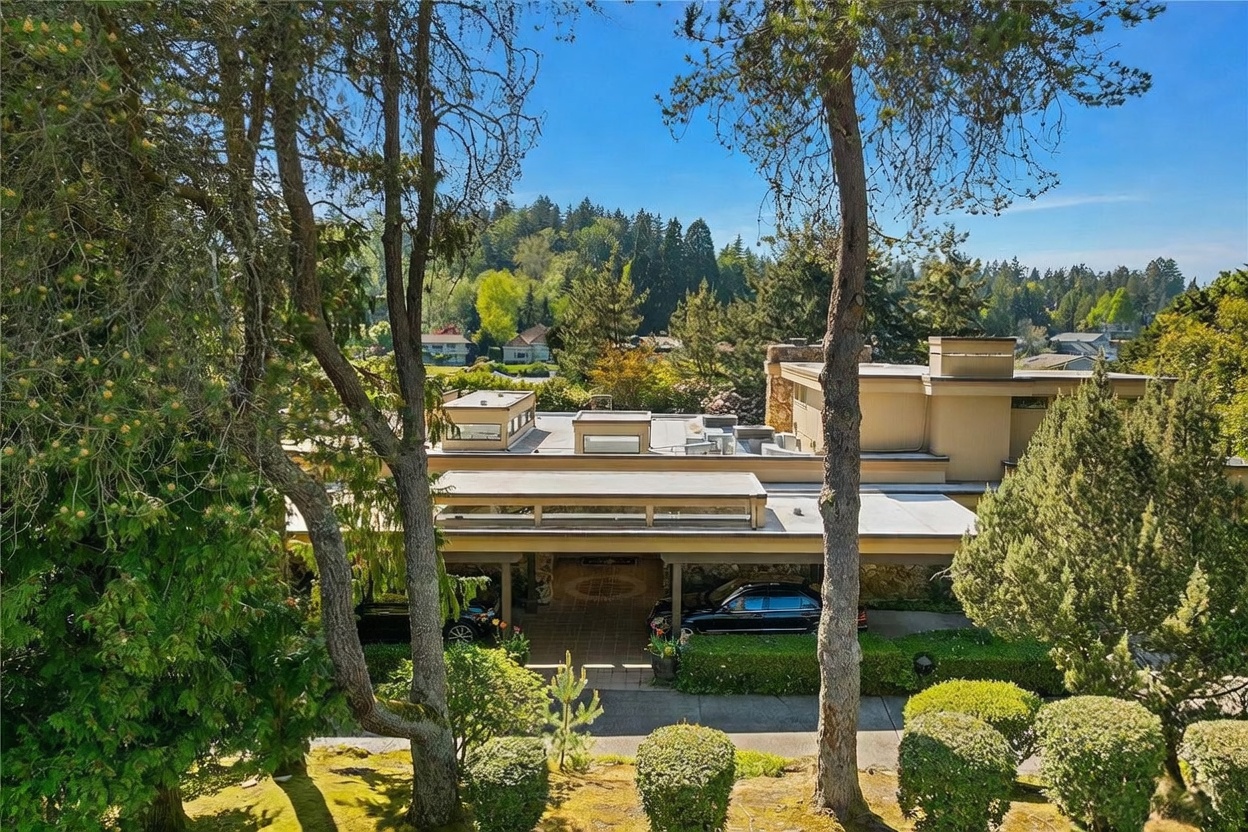 This elevated front view captures a mid-century modern residence nestled among mature trees and lush landscaping. The home features a flat roofline, horizontal siding, and a covered carport area that provides a sheltered entrance. The perspective highlights the architectural integration with the natural surroundings, showcasing a serene and private residential setting.