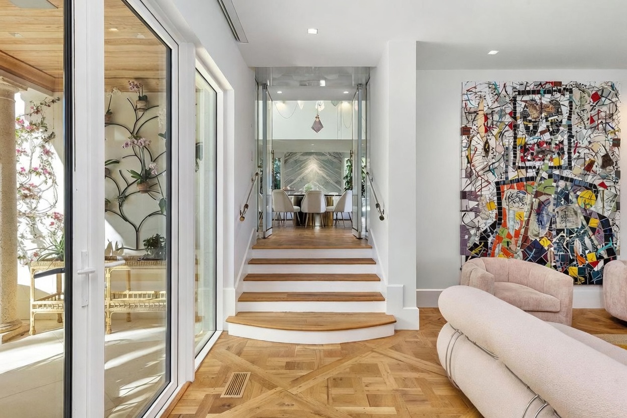 This elegant interior space features a set of wooden stairs leading up to a dining area framed by glass doors. To the right, a large, vibrant mosaic art piece hangs on the wall above a plush, light-colored sofa, while to the left, a glass wall offers a view into a bright, plant-filled atrium. The room is finished with intricate patterned wood flooring, creating a sophisticated and artistic atmosphere.