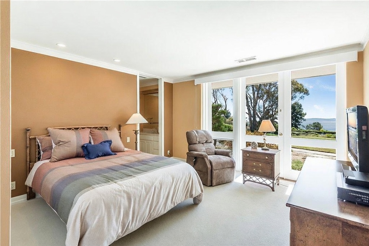 This bedroom features a neutral color palette with tan walls and light-colored carpeting, creating a calm and inviting atmosphere. A bed with a patterned headboard sits centrally, flanked by a lamp and a small nightstand, while a recliner is positioned near large windows that offer a scenic view of the outdoors. The room includes an open doorway leading to an en-suite bathroom and a television set on a dresser, providing a comfortable and functional living space.