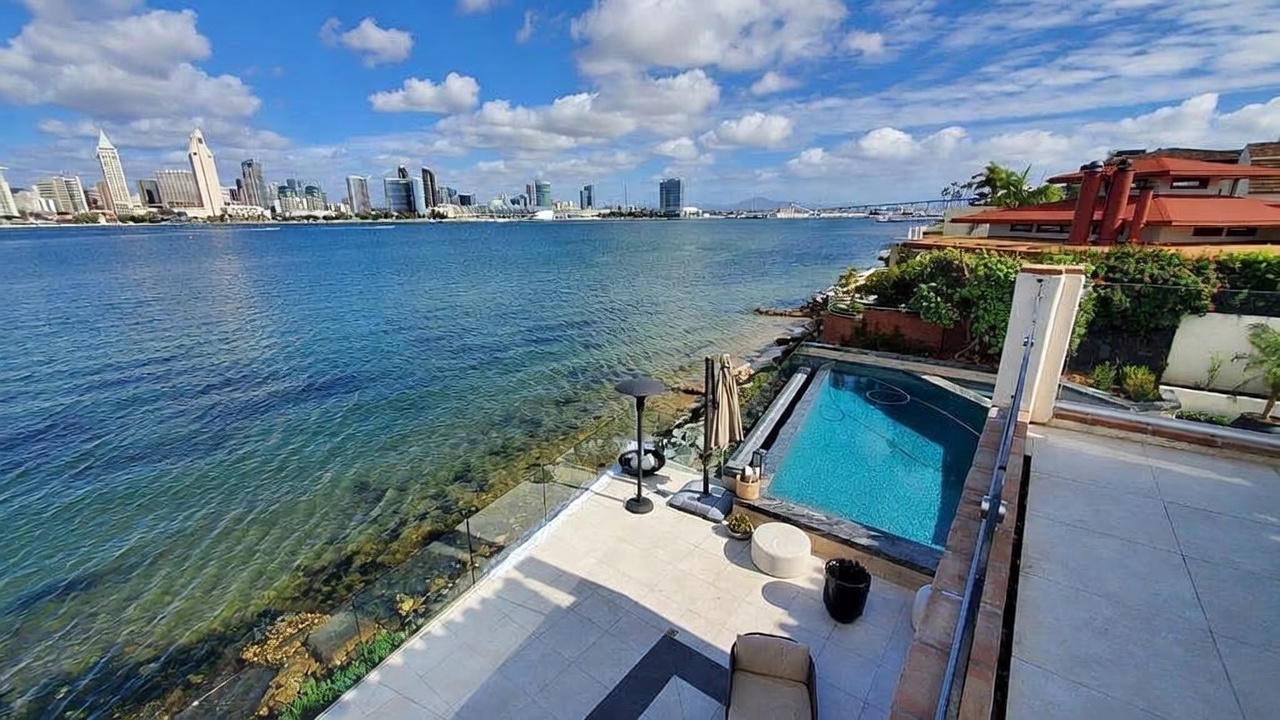 This elevated outdoor terrace offers a panoramic view of a bay and a distant city skyline under a bright, cloudy sky. The space features light-toned stone flooring, a glass-railed balcony, and a private plunge pool situated directly overlooking the water. A small seating area with a patio umbrella and a modern heater completes this luxurious waterfront retreat.