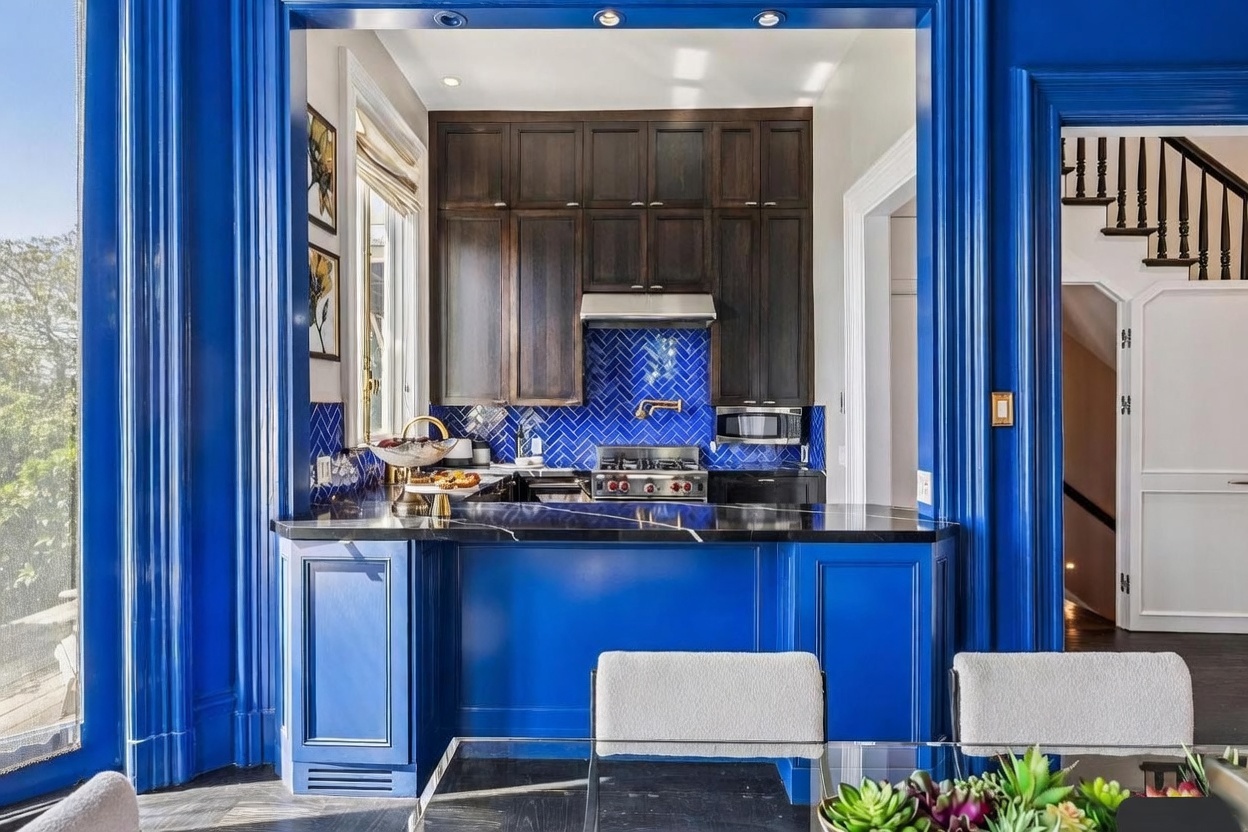 This vibrant kitchen features a bold, deep blue color palette contrasted by dark wood cabinetry and a striking herringbone-patterned blue tile backsplash. A sleek black stone countertop serves as a breakfast bar, overlooking a dining area with modern seating. The perspective is framed by a wide, blue-painted archway that creates a dramatic transition between the kitchen and the adjacent hallway and staircase.