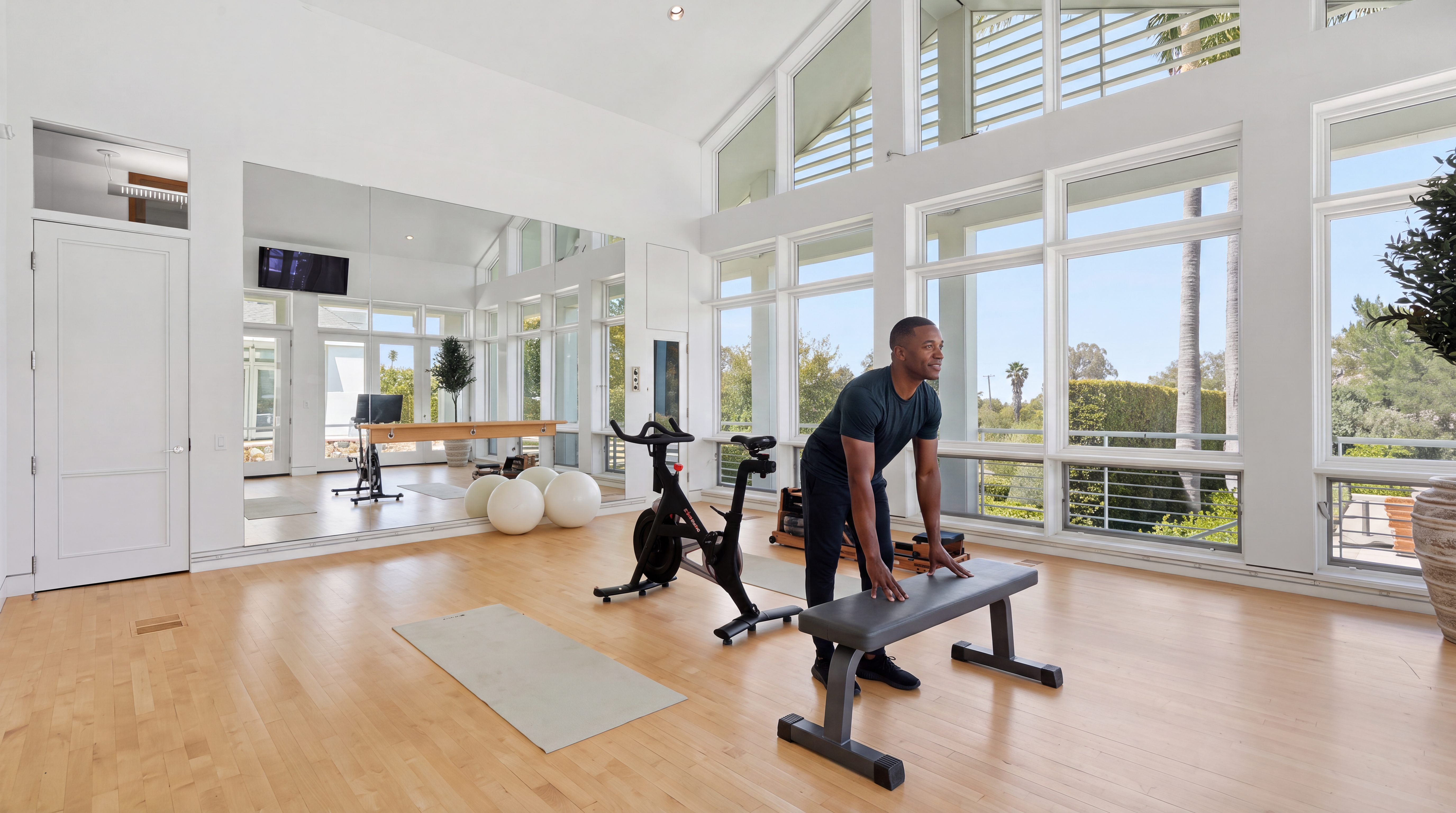 This spacious and light-filled home gym features high vaulted ceilings, expansive floor-to-ceiling windows, and warm hardwood flooring. It is equipped with a stationary bike, weight bench, exercise balls, and a pilates reformer, complemented by wall-to-wall mirrors. The room offers a serene and functional environment, perfect for a personal wellness routine with abundant natural light.