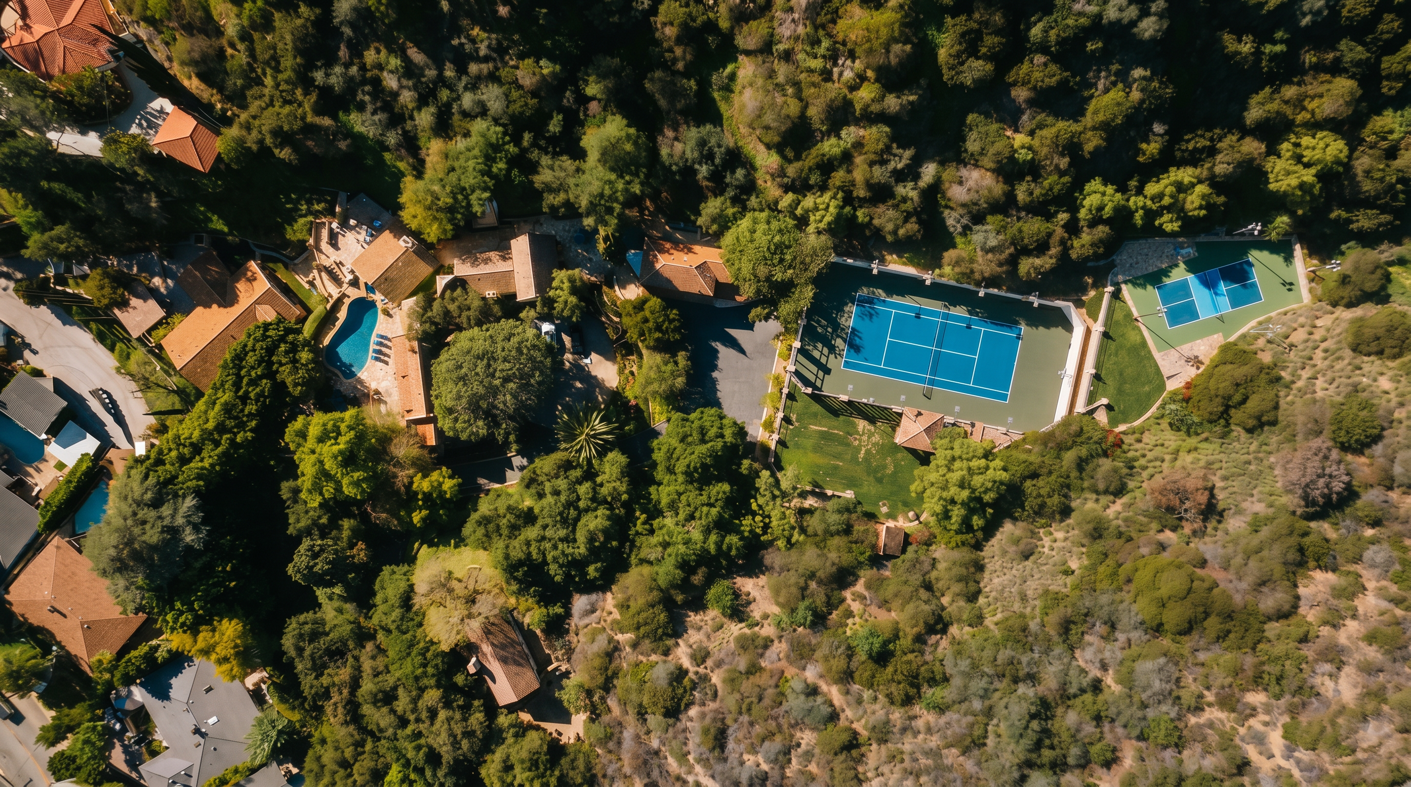 This high-angle aerial view captures a sprawling, secluded estate nestled within a dense, wooded hillside. The property features a large main residence with a swimming pool, a full-sized tennis court, and a secondary smaller court, all surrounded by lush, mature landscaping. The perspective emphasizes the expansive nature of the grounds and the private, tranquil setting of the home.