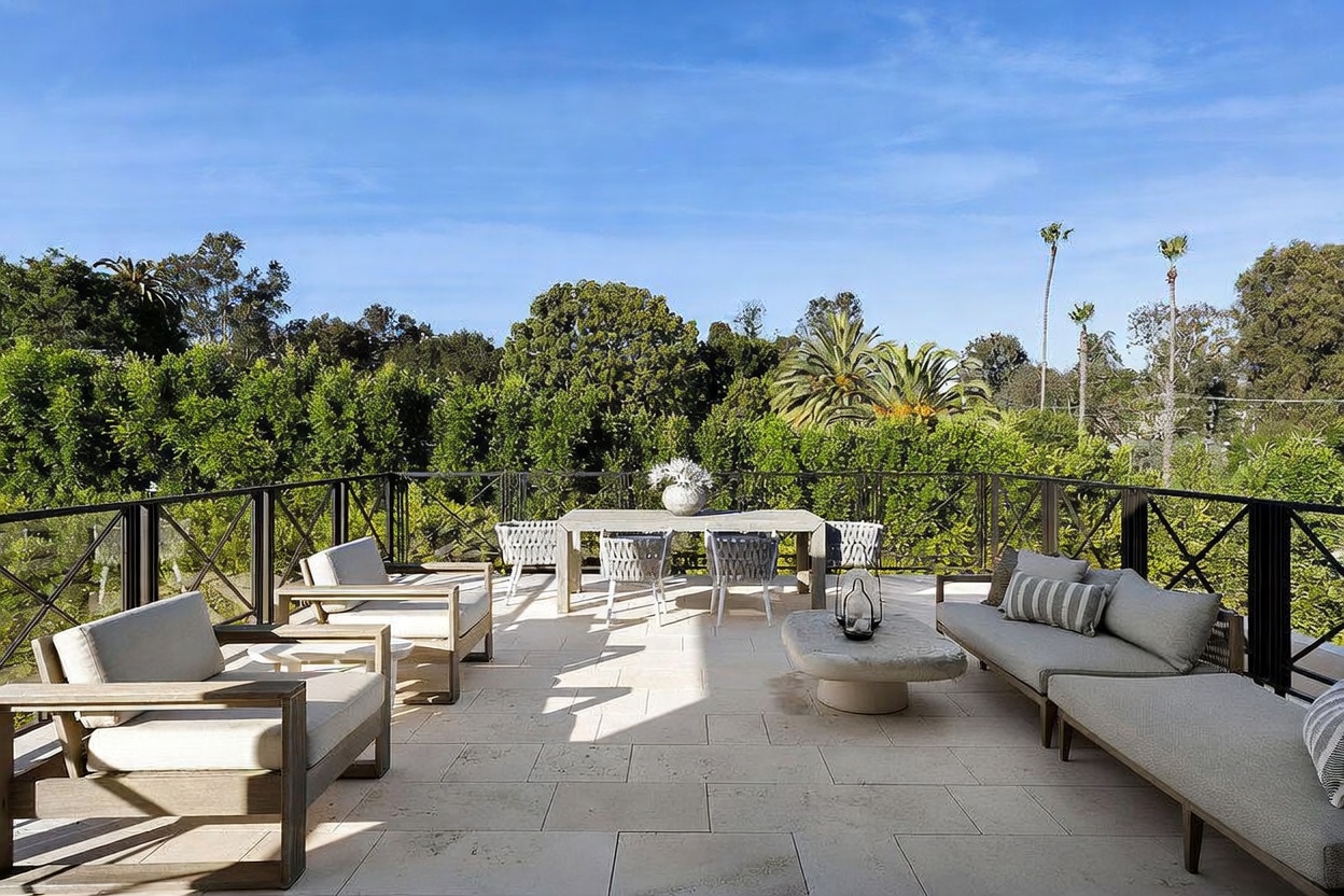 This expansive outdoor terrace features light-toned stone flooring and a sophisticated seating area arranged with modern, neutral-toned furniture. A dining table with woven chairs sits in the center, while a comfortable sofa and lounge chairs provide ample space for relaxation against a backdrop of lush green trees. The space is enclosed by a dark metal railing, offering an open-air atmosphere perfect for entertaining under the clear blue sky.
