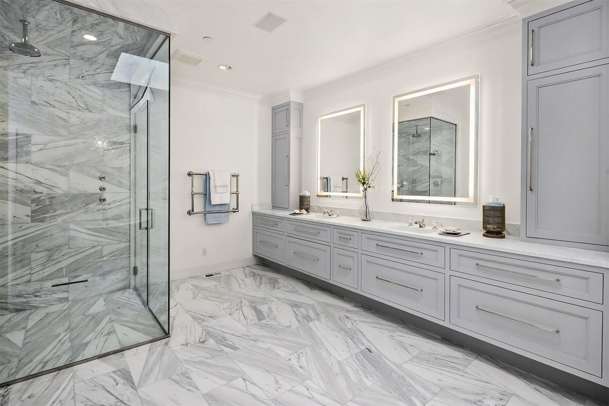 This spacious primary bathroom features a large walk-in shower with glass walls and patterned stone tile, paired with a long double vanity topped with light-colored stone. The room is illuminated by two large, rectangular mirrors with integrated LED lighting, creating a bright and modern atmosphere. The overall aesthetic is clean and sophisticated, with light gray cabinetry and matching stone flooring throughout.