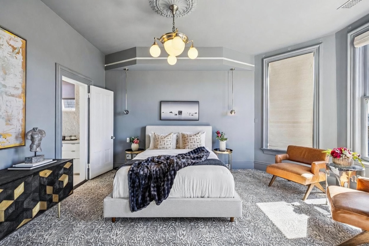This elegant primary bedroom features a central bed with a neutral upholstered headboard, accented by a dark, textured throw blanket and patterned pillows. The room is framed by a soft blue wall color, a decorative ceiling medallion with a brass chandelier, and a striking black and gold geometric dresser. Large windows allow natural light to illuminate the patterned carpet, while two leather armchairs provide a comfortable seating area near the window.