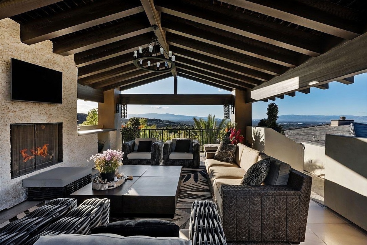 This expansive covered outdoor living space features a vaulted ceiling with exposed dark wood beams and a central chandelier. A stone-clad chimney breast houses a fireplace, while a large sectional sofa and coffee table create a comfortable seating area. The space opens to a panoramic view of the surrounding landscape and distant mountains, framed by a metal railing.