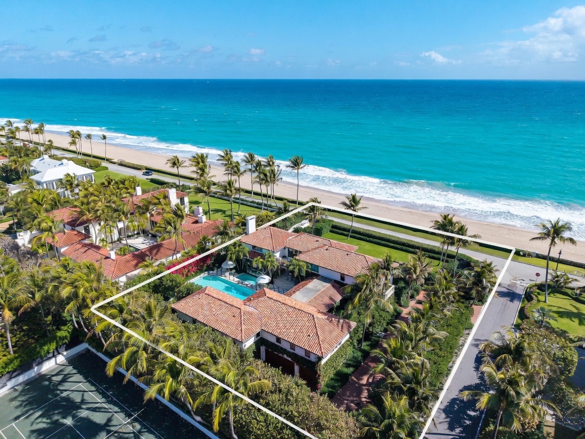 This high-angle aerial view captures a sprawling, Mediterranean-style estate situated directly along a pristine coastline. The property features a central courtyard with a swimming pool, surrounded by lush tropical landscaping and a terracotta-tiled roof. A white boundary line highlights the expansive lot, which sits adjacent to a quiet coastal road and the turquoise waters of the ocean.