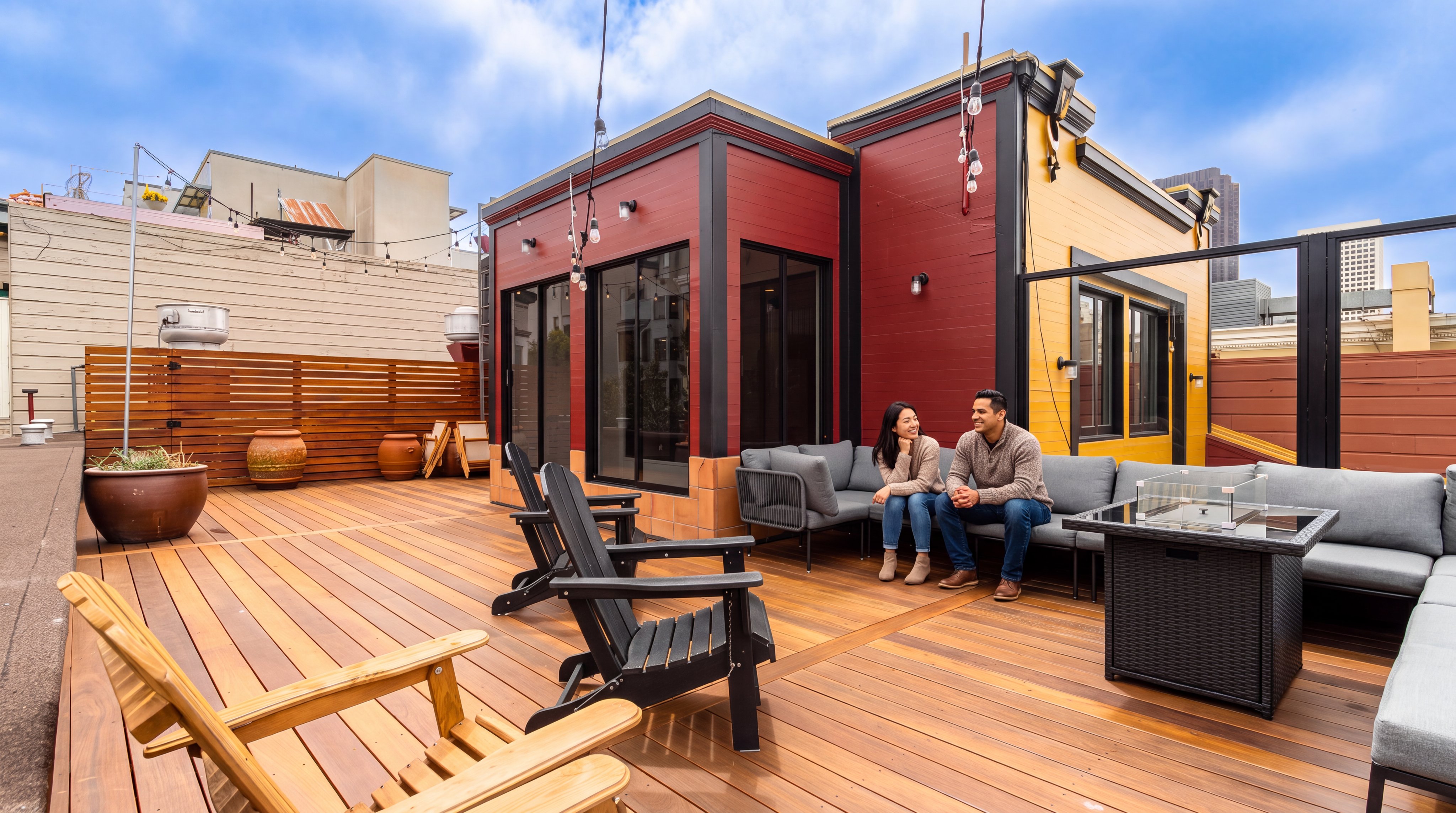 This spacious rooftop deck features high-quality natural wood decking, modern outdoor sofa seating, and classic adirondack chairs, creating an inviting entertainment area. The structure displays a contemporary aesthetic with bold red and yellow exterior walls and large sliding glass doors, seamlessly blending indoor and outdoor living. The setting is ideal for socializing, accented by string lights and a panoramic urban skyline backdrop.