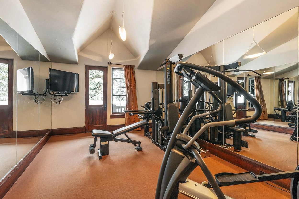 This home gym features a vaulted ceiling and a wall of mirrors that enhances the sense of space. The room is equipped with a multi-station weight machine, a weight bench, and an elliptical trainer, all set against a neutral carpeted floor. A door with glass panels and a window with brown curtains provide natural light, while a wall-mounted television offers entertainment during workouts.