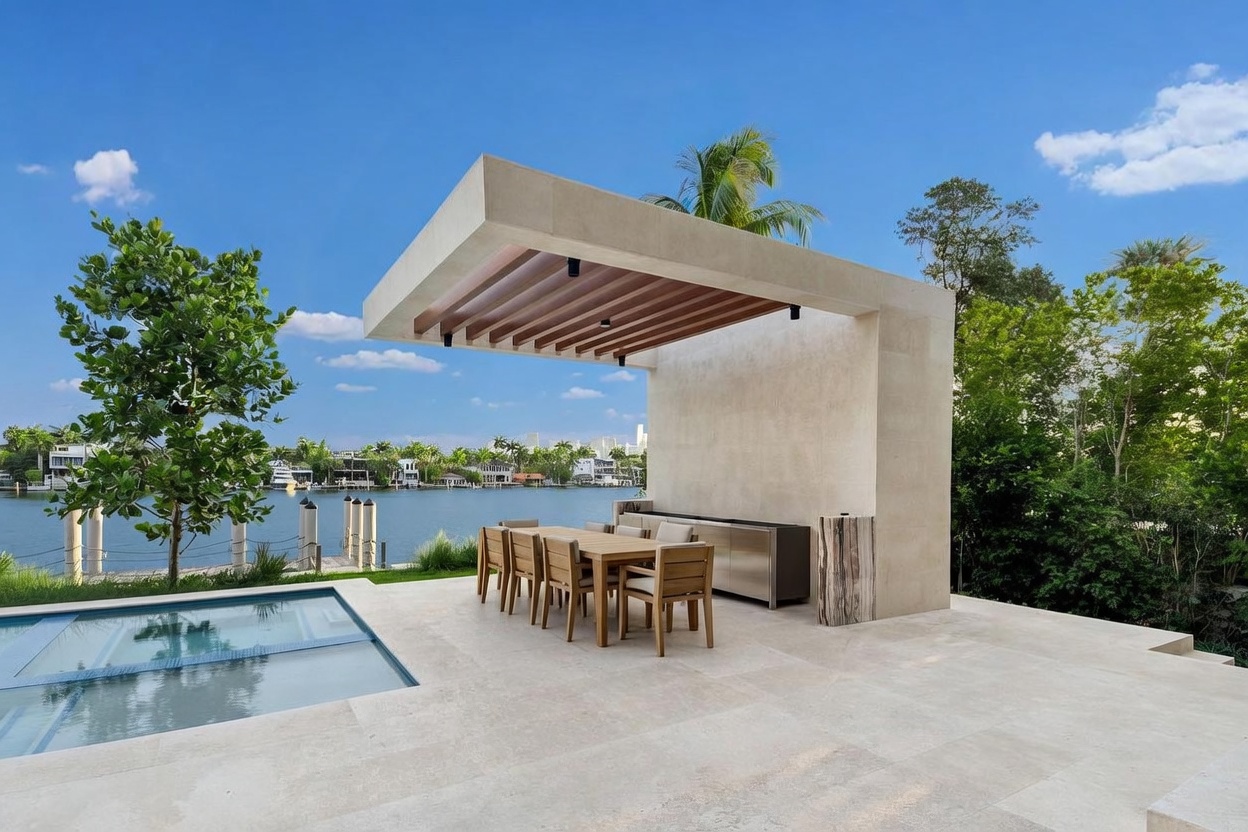 This luxurious outdoor living space features a modern, covered dining area with a sleek, minimalist pergola structure overlooking a waterfront view. A pristine swimming pool with a glass-paneled edge sits adjacent to the stone-paved patio, which is furnished with a wooden dining set. The scene captures a serene, high-end waterfront lifestyle with lush greenery and a clear blue sky in the background.