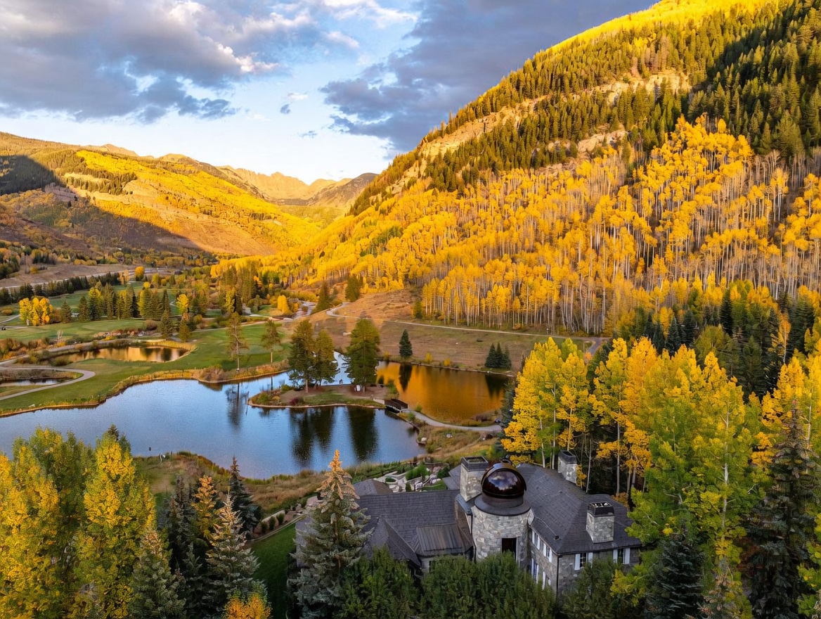 This high-angle aerial shot captures a luxurious stone estate nestled within a vibrant autumn landscape of golden aspen trees and evergreen forests. The property features a distinctive domed observatory tower and overlooks a serene, reflective pond set against a backdrop of dramatic mountain peaks. The golden hour light enhances the warm, seasonal colors, creating a picturesque and tranquil mountain retreat atmosphere.