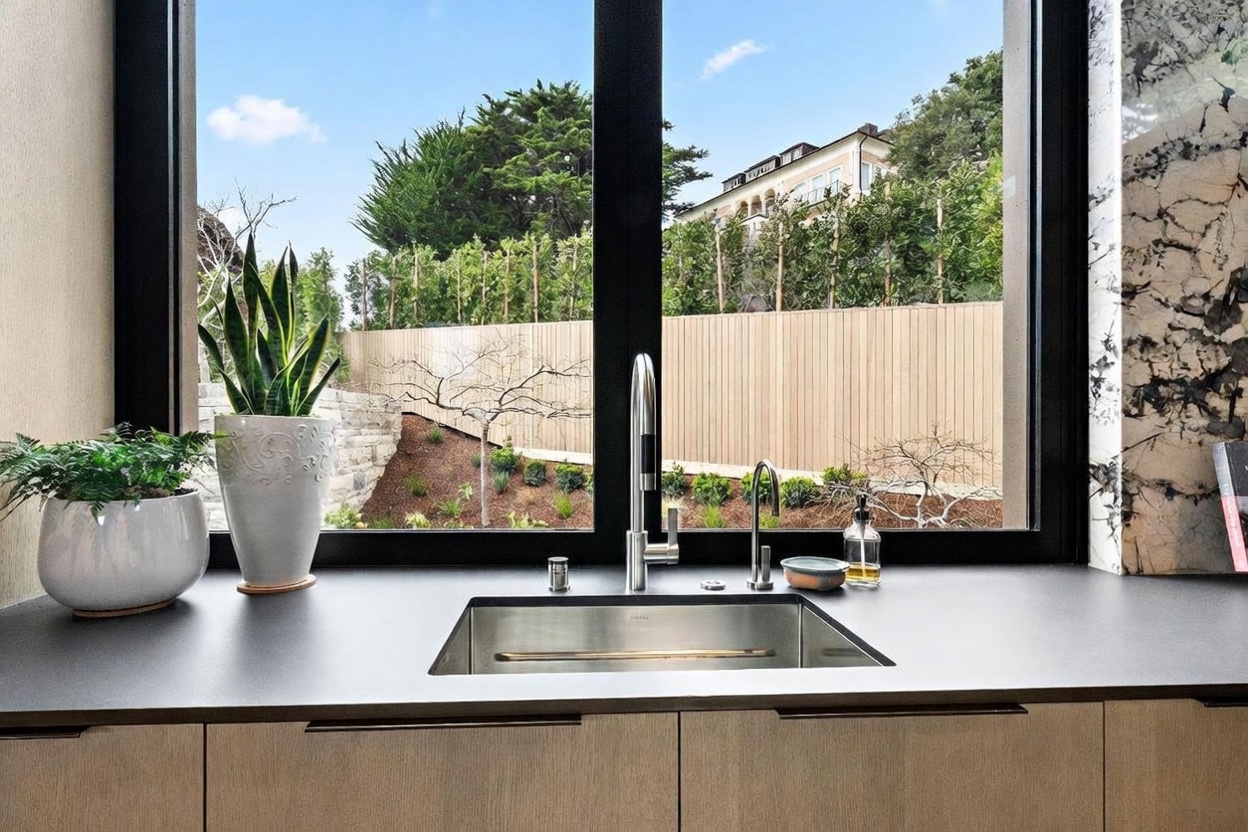 This interior shot features a sleek, modern kitchen sink area positioned directly in front of a large window that overlooks a landscaped backyard. The dark countertop contrasts with the light wood-toned cabinetry below, while a stainless steel sink and contemporary faucet fixtures provide a clean, functional aesthetic. Two potted plants sit on the counter, adding a touch of greenery that complements the outdoor view.