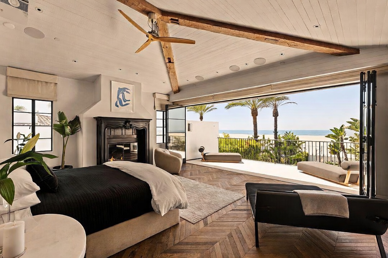This luxurious primary bedroom features a sophisticated design with dark herringbone wood flooring and a prominent black fireplace surround. Large, retractable glass walls open the space to a private balcony, offering an expansive, unobstructed view of the ocean and beach. The room is accented by a wooden ceiling beam, a modern ceiling fan, and minimalist furniture, creating an airy and high-end coastal atmosphere.