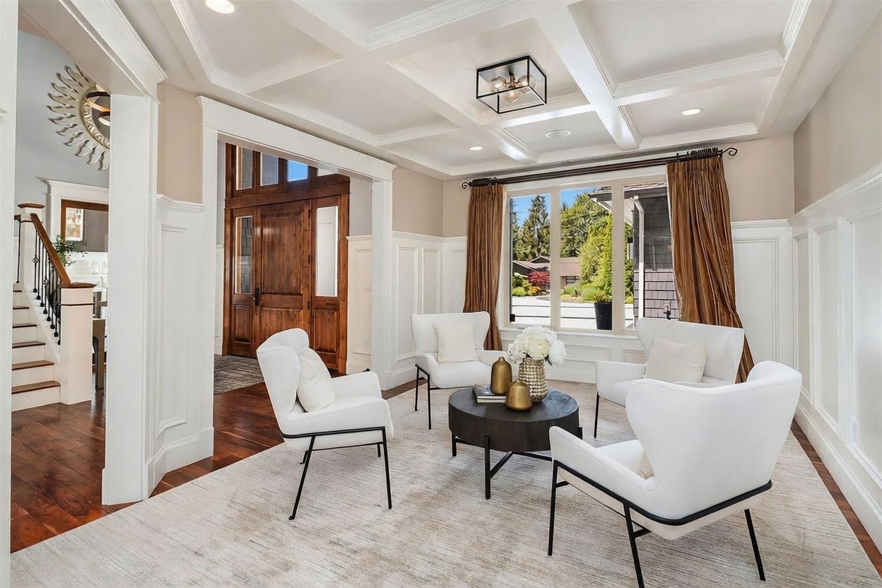 This elegant living room features a coffered ceiling and crisp white wainscoting, creating a sophisticated and airy atmosphere. The space is furnished with four modern white armchairs arranged around a dark circular coffee table, all set upon a textured neutral area rug. Large windows draped in warm-toned curtains allow natural light to flood the room, while a view into the adjacent entryway reveals a grand wooden door and a staircase.