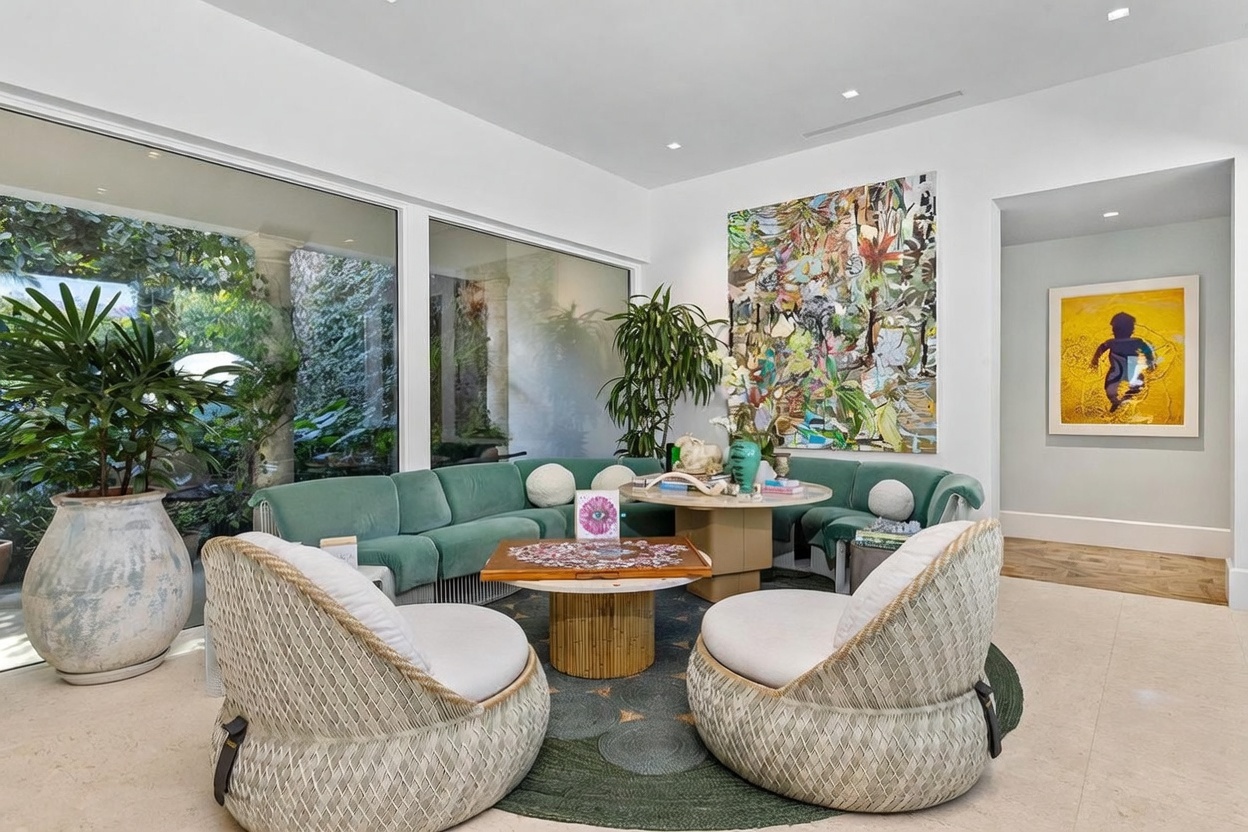 This bright and airy living space features a large, curved teal sofa arranged around a central coffee table, complemented by two textured, rounded armchairs. The room is anchored by a circular rug and showcases a large, vibrant abstract painting on the wall, alongside a smaller framed piece in an adjacent hallway. Large windows provide an expansive view of the lush, green outdoor foliage, creating a seamless connection between the interior and the garden.