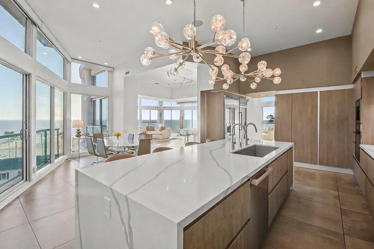 This bright, open-concept kitchen features a large central island with a stone countertop and light wood cabinetry. A modern, multi-bulb chandelier hangs above the island, while large windows provide expansive views of the ocean in the background. The space flows seamlessly into a dining area and living room, creating an airy and sophisticated atmosphere.