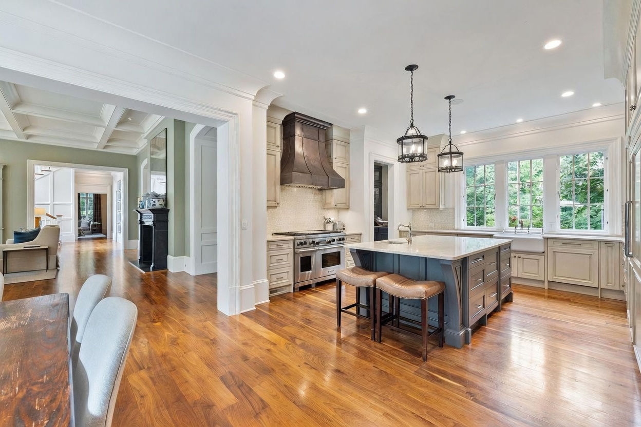 This spacious, open-concept kitchen features a large central island with bar seating, dark cabinetry, and light-toned stone countertops. A prominent metal range hood serves as a focal point above the professional-grade stove, while tall windows above the farmhouse sink provide ample natural light. The warm wood flooring flows seamlessly into an adjacent living area, creating an inviting and cohesive living space.