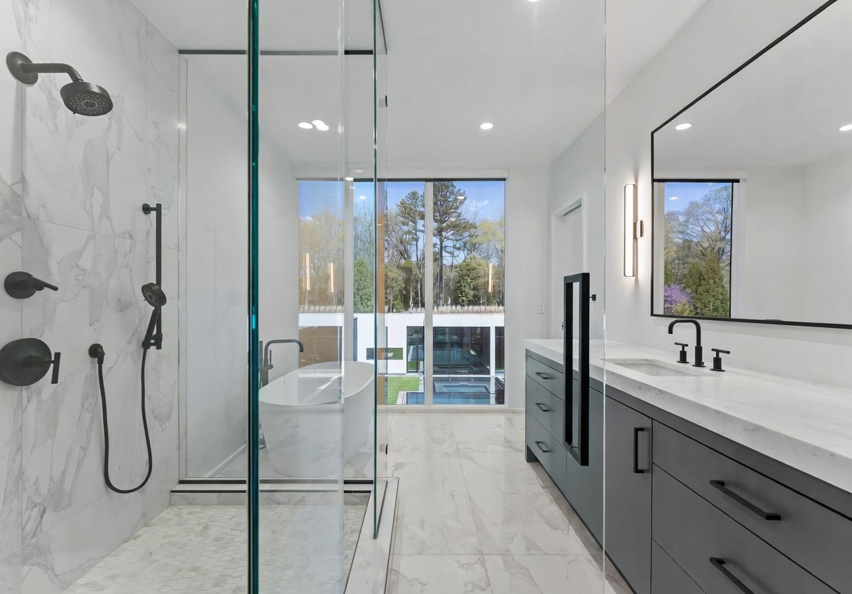 This modern primary bathroom features a sleek glass-enclosed walk-in shower with patterned tile flooring and a dark gray vanity with a light-toned stone countertop. A freestanding soaking tub is positioned in front of large windows that provide a clear view of the outdoor pool area. The space is defined by its clean lines, minimalist aesthetic, and bright, airy atmosphere.