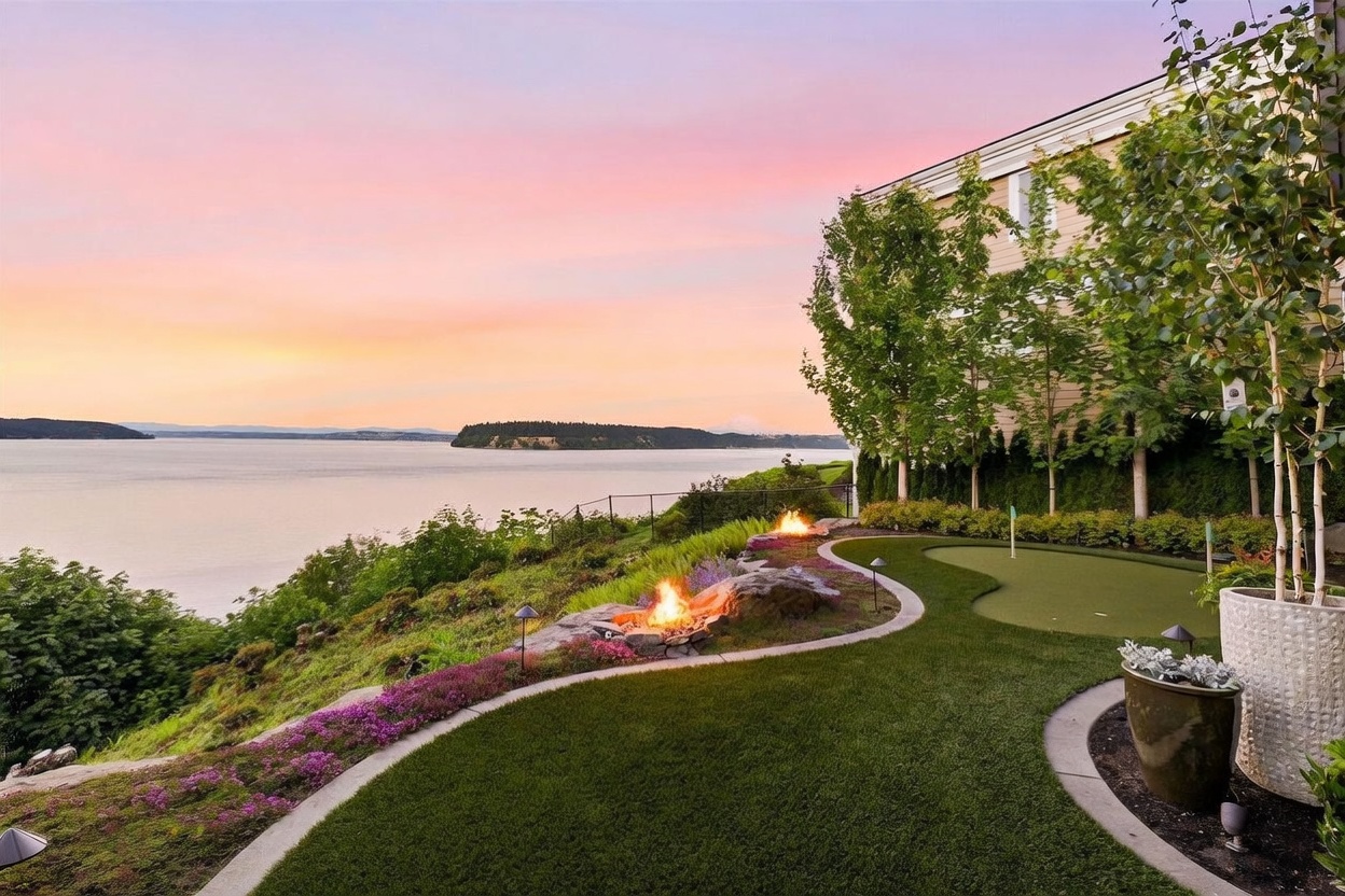 This stunning backyard features a manicured putting green and a stone-lined fire pit area, perfectly positioned to overlook a vast, tranquil body of water. The scene is captured at sunset, with a vibrant pink and orange sky casting a warm glow over the lush lawn, mature trees, and decorative purple flowers. The perspective highlights the seamless integration of luxury outdoor living space with the natural beauty of the waterfront landscape.