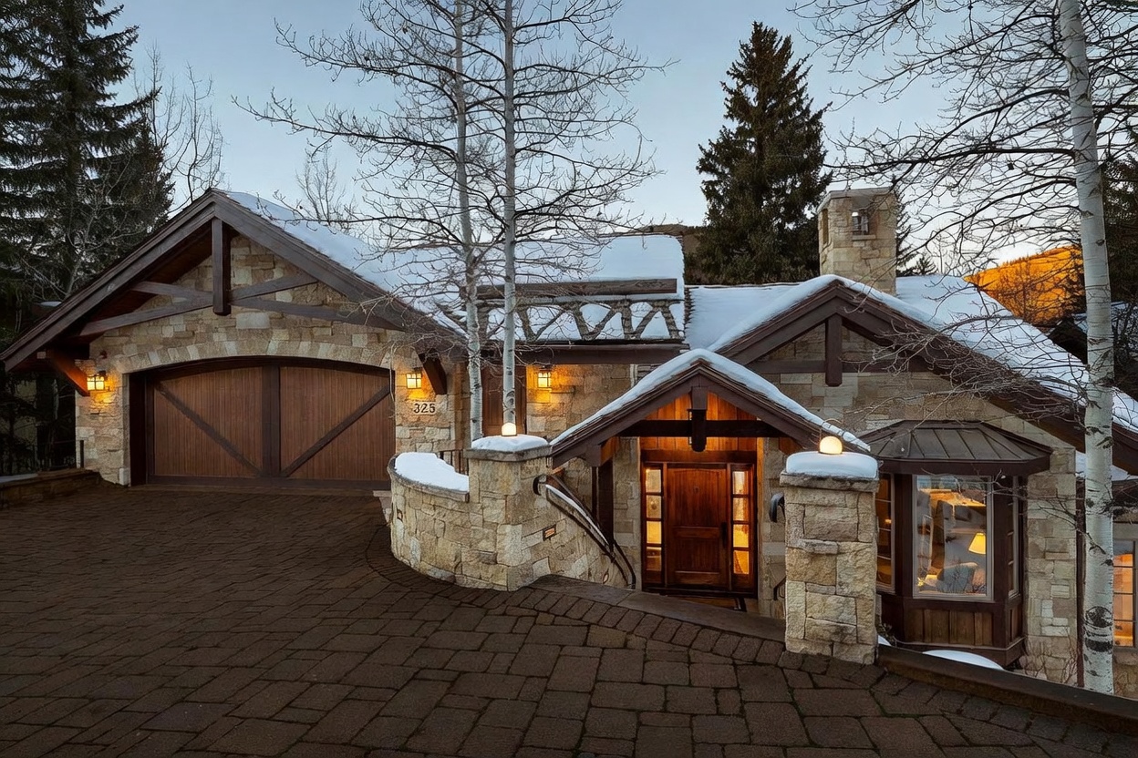 This mountain-style residence features a striking exterior clad in textured stone with dark wood trim and a prominent gabled roofline. The front entrance is accessed via a stone-paved driveway leading to a recessed wooden door, flanked by stone pillars and a bay window. The scene is set in a snowy, alpine environment with bare aspen trees, creating a cozy and upscale winter retreat aesthetic.