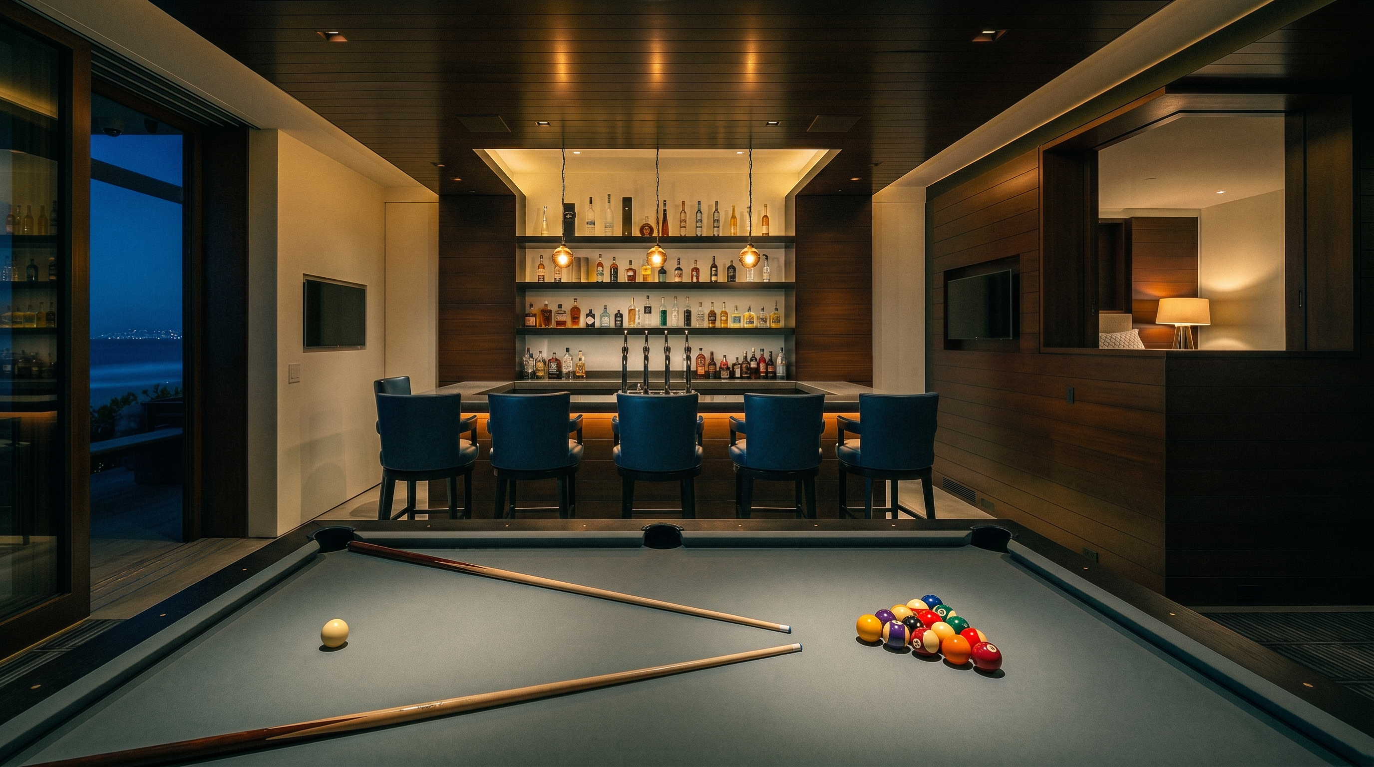 This sophisticated game room features a central pool table with a grey felt surface and a custom-built bar area with dark wood paneling and integrated shelving. Five blue leather bar stools are arranged along the bar, which is illuminated by three pendant lights hanging from the wood-paneled ceiling. Large sliding glass doors on the left provide a direct view of the ocean and beach, creating a seamless indoor-outdoor connection.