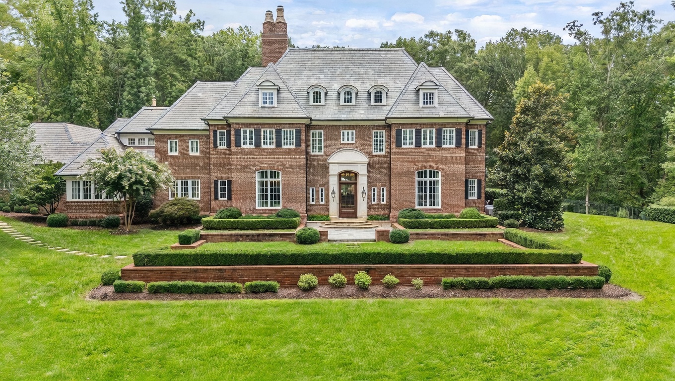 This grand, multi-story brick estate features a symmetrical Georgian-inspired facade with a prominent central entryway and a steep, slate-tiled roof punctuated by multiple dormers. The property is set against a lush, mature forest backdrop and includes a meticulously landscaped tiered garden with manicured boxwood hedges and brick retaining walls. The perspective is a straight-on, eye-level shot that captures the stately architecture and the expansive, well-maintained front lawn.