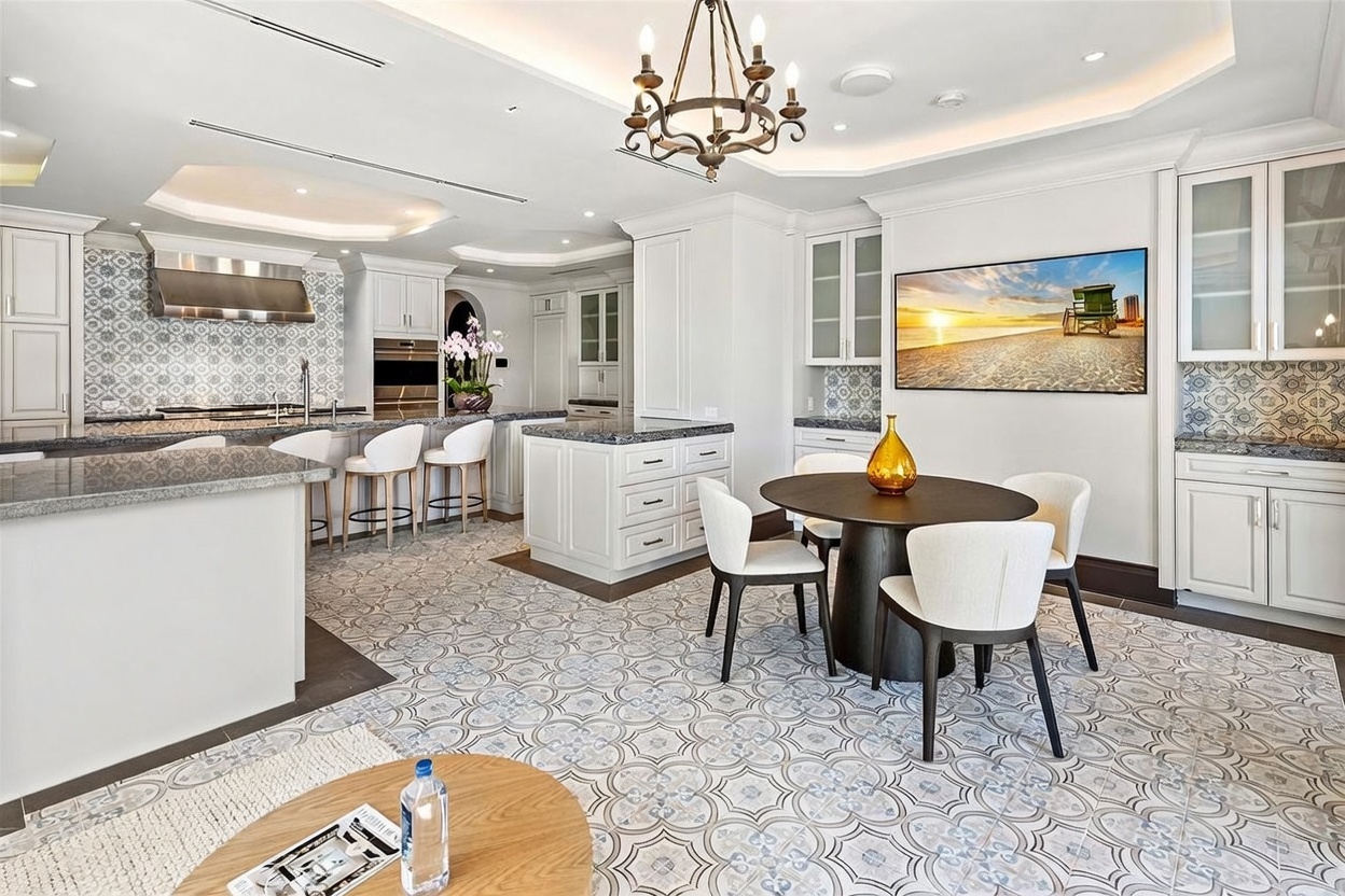This bright and spacious kitchen features white cabinetry, a large central island with stone countertops, and a breakfast nook with a round table. The space is defined by intricate patterned floor tiles, a decorative backsplash, and a classic chandelier hanging above the dining area. A large television is mounted on the wall, and the open-concept layout provides a seamless flow between the cooking and dining zones.