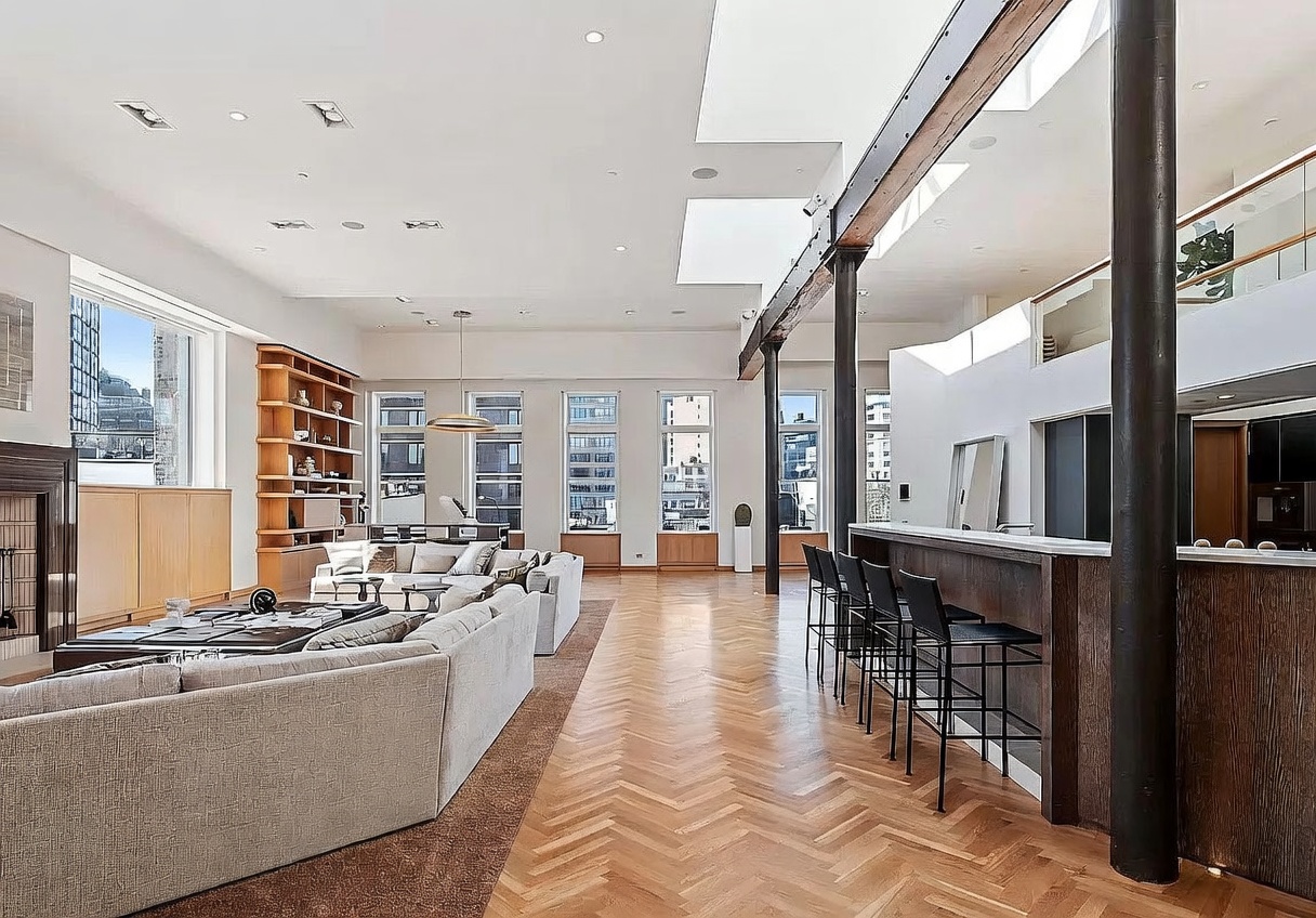 This expansive, open-concept living area features a high ceiling with exposed structural beams and skylights that flood the space with natural light. The room is anchored by a large, neutral-toned sectional sofa and a tall fireplace surround, complemented by herringbone-patterned wood flooring throughout. A long bar counter with stools separates the living space from the kitchen area, creating a sophisticated and airy atmosphere suitable for entertaining.