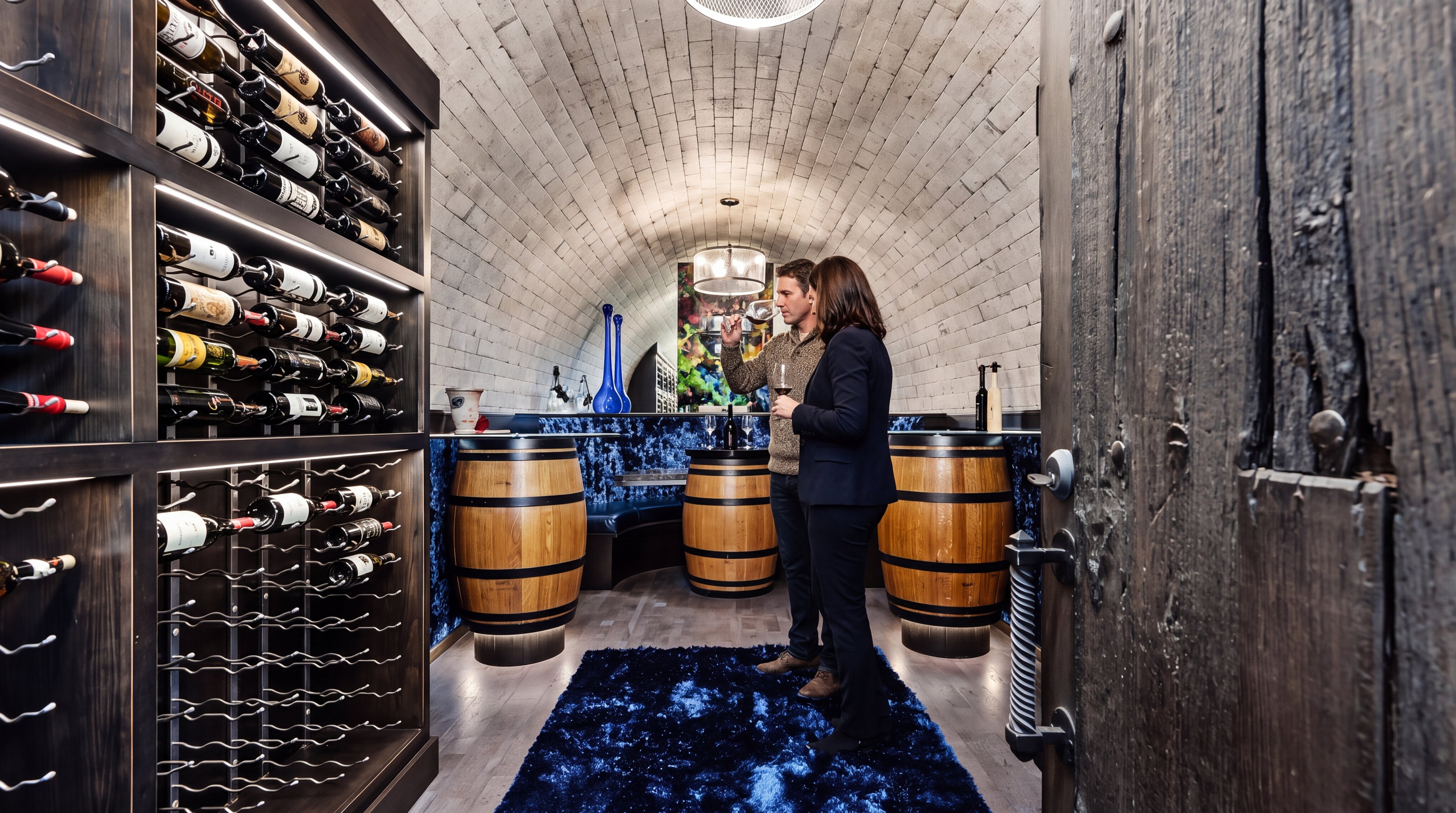 This luxurious wine cellar features a dramatic vaulted stone ceiling, custom dark wood shelving, and integrated LED lighting. A blue shag area rug adds a pop of color, while repurposed wine barrels serve as tasting tables, creating an intimate and sophisticated atmosphere perfect for wine enthusiasts.