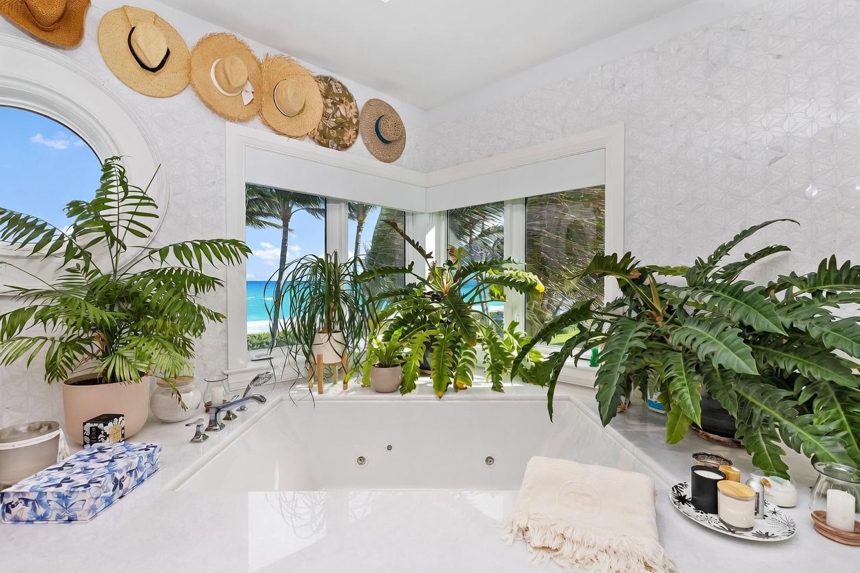 This bright and airy primary bathroom features a large soaking tub positioned in a corner with expansive windows that offer a stunning, direct view of the ocean and palm trees. The space is decorated with a collection of straw hats mounted on the wall and lush potted plants, creating a relaxed, tropical spa-like atmosphere. The clean, white aesthetic is complemented by patterned wall tiles and a marble-look deck surrounding the tub.