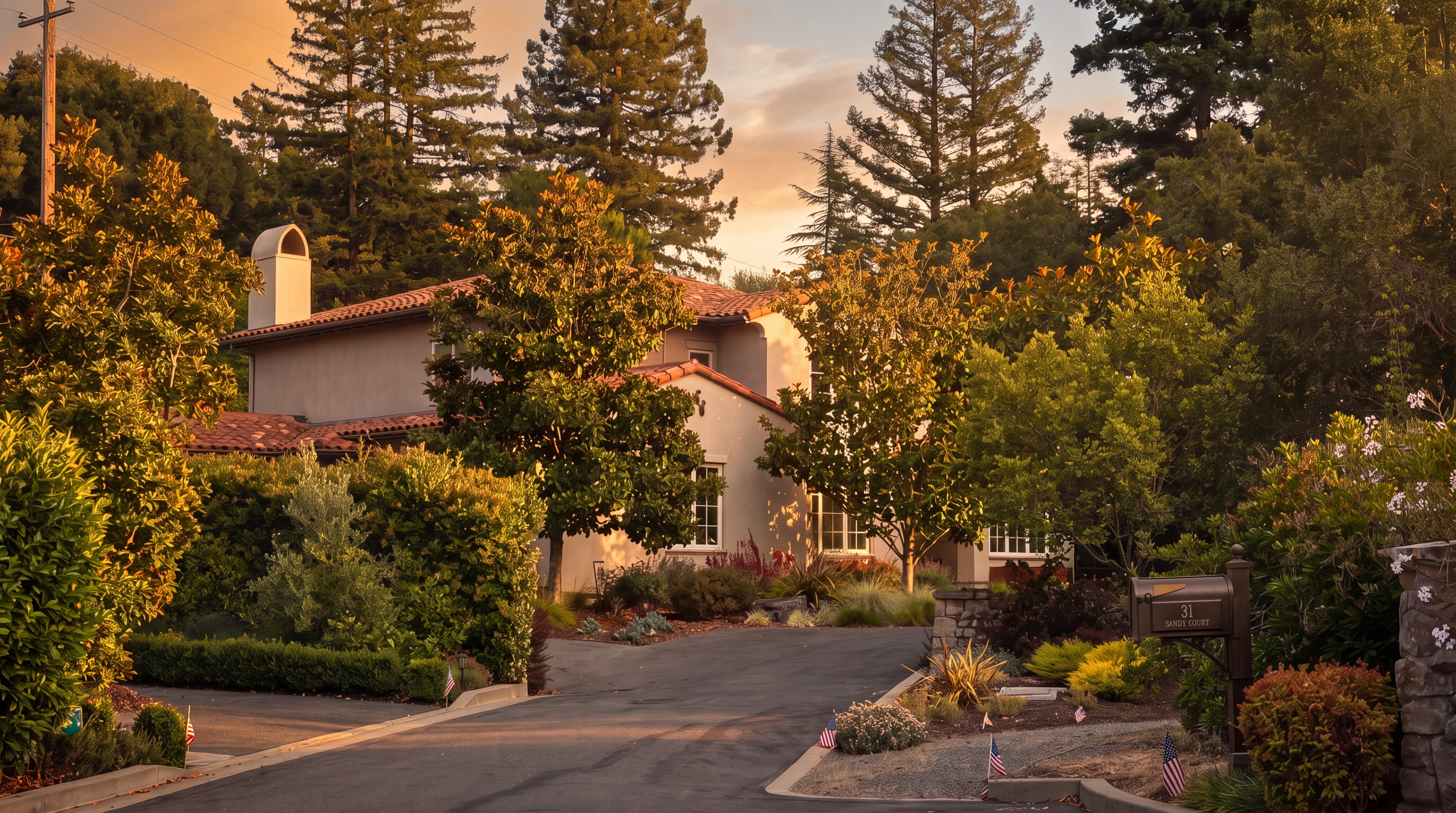 This front-facing view captures a multi-story residence with a classic Mediterranean-inspired aesthetic, featuring a terracotta tile roof and a light-toned stucco facade. The home is nestled among mature trees and lush landscaping, with a paved driveway leading up to the entrance. A mailbox marked '31 Sandy Court' stands in the foreground, and the scene is framed by a bright blue sky, creating a welcoming and private atmosphere.