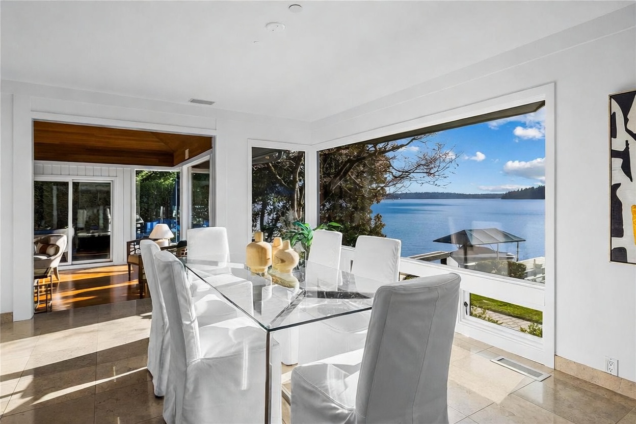 This bright and airy dining room features a modern glass-top table surrounded by white slipcovered chairs, creating a clean and sophisticated aesthetic. A large, expansive window provides a stunning, panoramic view of the serene lake and surrounding trees, flooding the space with natural light. The room is finished with polished tile flooring and flows seamlessly into an adjacent sunlit living area, emphasizing an open and inviting atmosphere.