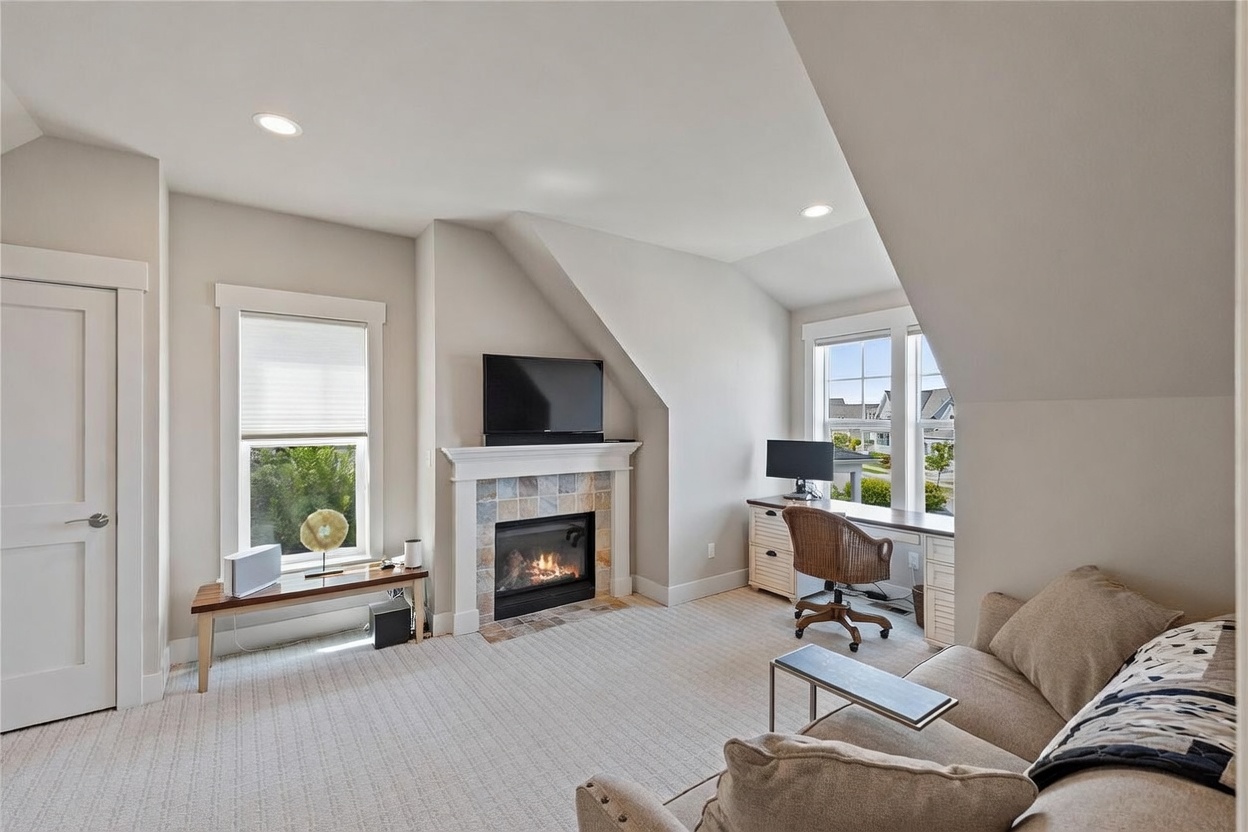 This cozy, multi-functional living space features a neutral color palette, plush carpeting, and a central fireplace with a decorative tile surround and a white mantel. To the right, a built-in desk area provides a dedicated workspace, while a comfortable sofa sits in the foreground, creating a relaxed atmosphere. The room is illuminated by recessed lighting and natural light streaming through two windows, offering a view of the neighborhood.
