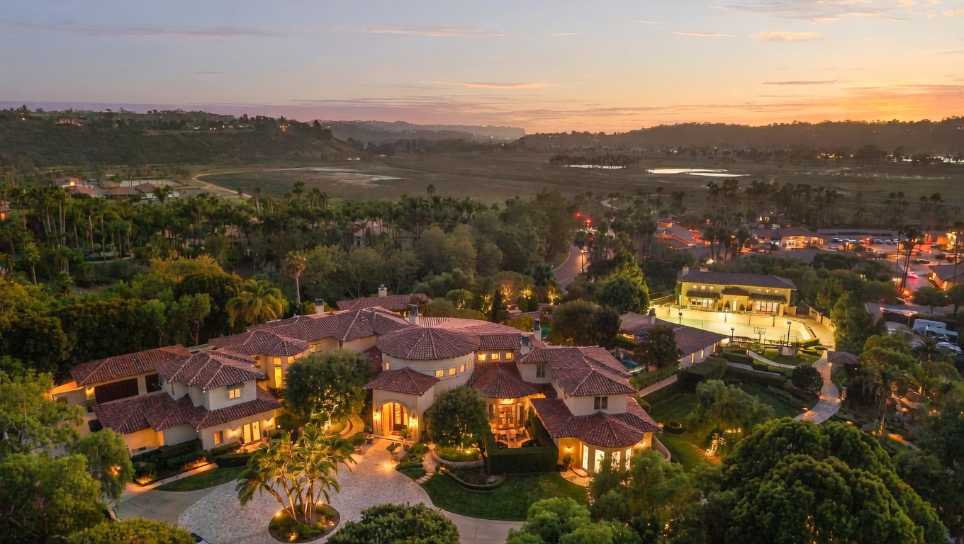 This aerial view showcases a sprawling estate with a terracotta-tiled roof, surrounded by lush greenery and mature trees. The property features a circular driveway and is nestled in a scenic landscape with a view of a marsh and distant hills. The overall impression is one of luxury, privacy, and tranquility.