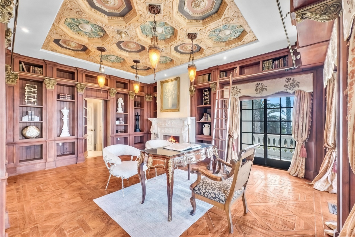 This elegant home office features intricate wood paneling, built-in bookshelves, and a highly detailed coffered ceiling with decorative molding. A central desk sits on a light-colored rug, complemented by a white accent chair and a patterned upholstered chair, while a rolling library ladder provides access to the upper shelves. The room is anchored by a stone fireplace and large glass doors that open to a balcony, creating a sophisticated and scholarly atmosphere.