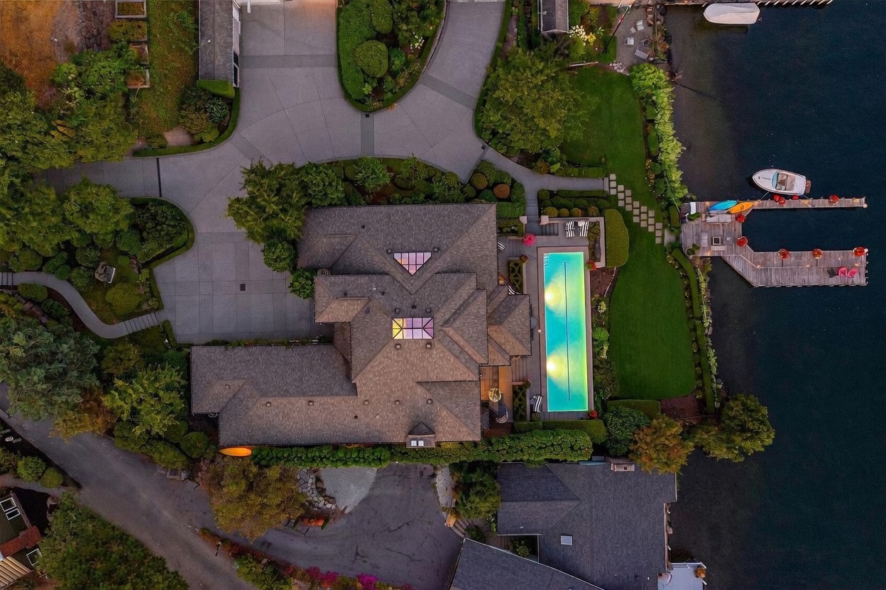This high-angle aerial perspective captures a luxurious waterfront estate at dusk, showcasing a sprawling residence with a complex shingled roof and two prominent skylights. The property features a rectangular swimming pool illuminated from within, a manicured lawn, and a private wooden dock extending into the dark water, where a small boat is moored. The scene is framed by lush landscaping, a paved driveway, and neighboring homes, creating an impression of exclusive, serene lakeside living.