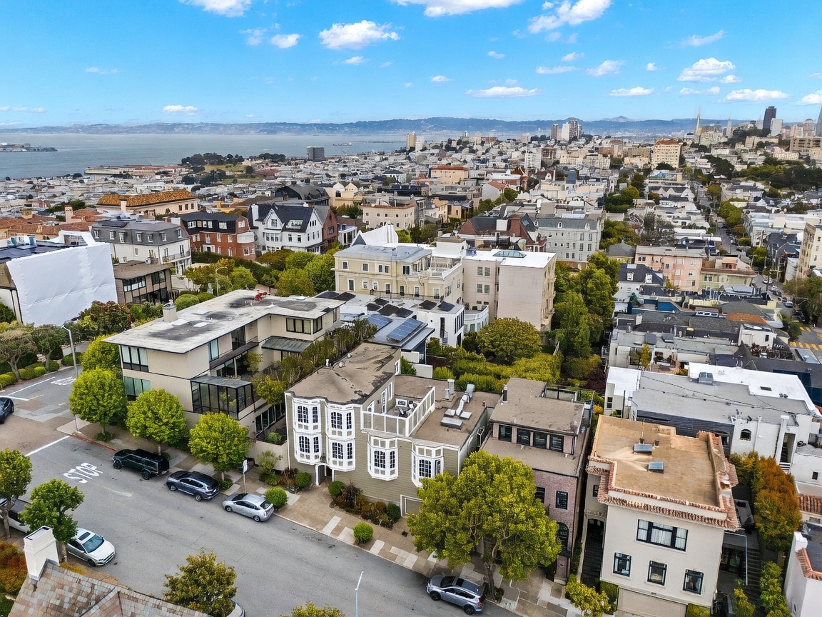 This high-angle aerial view captures a dense urban neighborhood in San Francisco, showcasing a mix of multi-story residential buildings and townhomes. The perspective highlights the proximity to the bay and the city skyline in the distance, emphasizing the prime location and architectural density of the area. The scene is bathed in bright daylight, revealing tree-lined streets and the characteristic terraced layout of the city's housing.