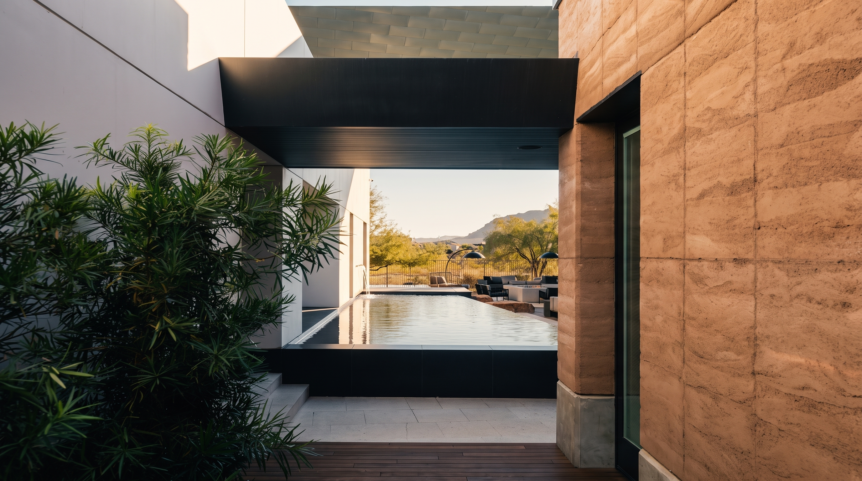 This modern architectural space features a sleek, rectangular lap pool framed by a dark, covered walkway that creates a dramatic transition between the indoor and outdoor areas. To the right, a textured, earth-toned wall adds warmth, while a lush green plant in the foreground provides a soft contrast to the clean lines of the structure. The view through the covered passage reveals a desert landscape with distant mountains, emphasizing a seamless connection between luxury living and the natural environment.