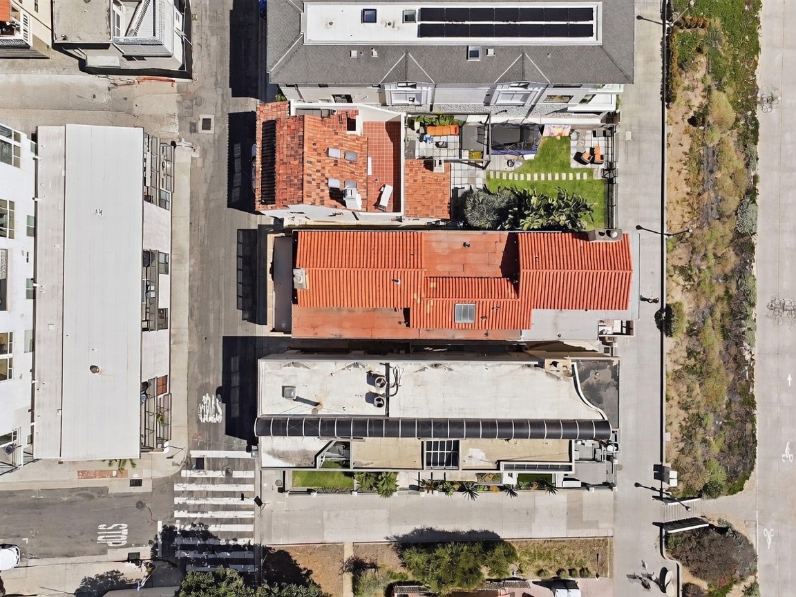 This top-down aerial view captures a dense urban residential block, showcasing a row of homes with distinct architectural styles and rooflines. The perspective highlights the layout of the properties, including a house with a prominent red-tiled roof, a neighboring home with a green lawn and patio, and a flat-roofed structure with solar panels. The scene is framed by a street with a crosswalk on one side and a natural, landscaped area on the other, providing a clear sense of the property's location and neighborhood density.