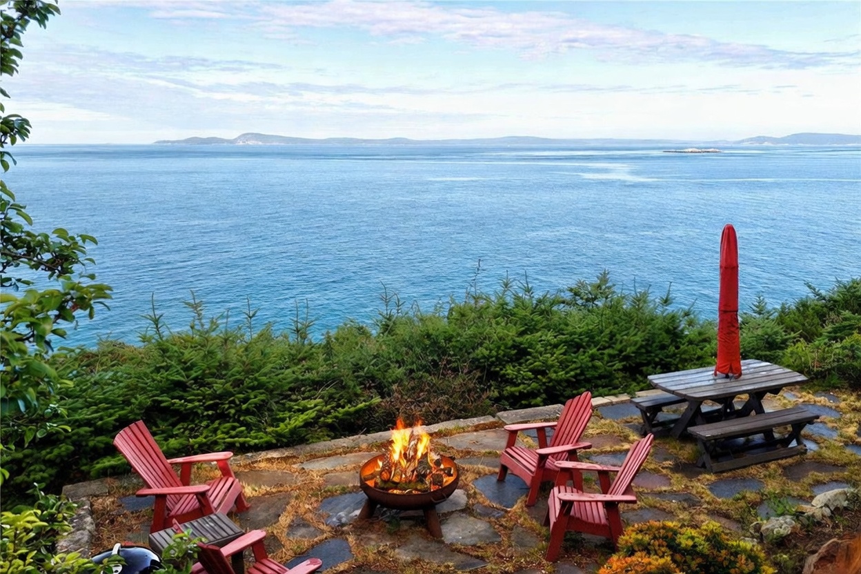 This scenic outdoor patio features a stone-paved seating area centered around a metal fire pit, surrounded by four red Adirondack chairs. A wooden picnic table with a closed red umbrella sits to the side, all overlooking a vast, tranquil ocean view with distant islands on the horizon. The space is framed by lush evergreen trees, creating a peaceful and private atmosphere perfect for relaxation.
