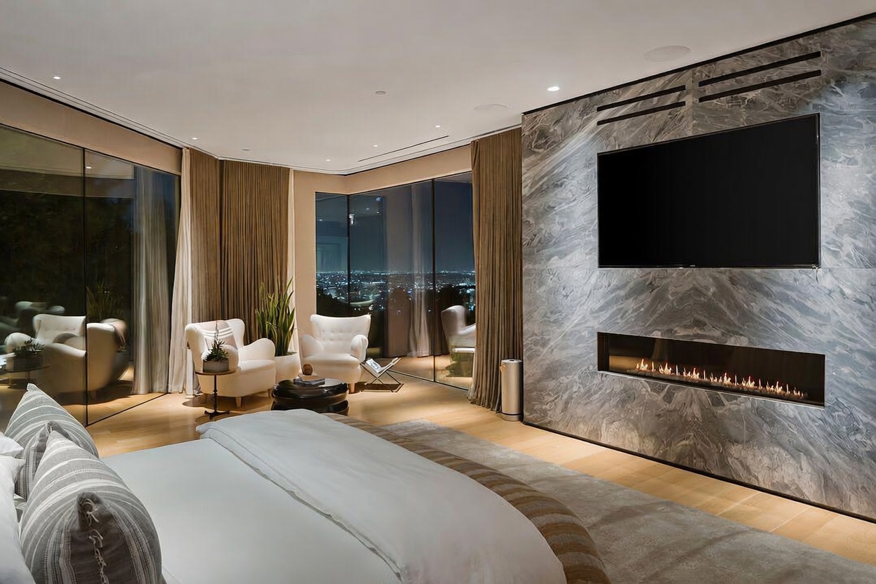 This luxurious primary bedroom features a large bed in the foreground and a striking stone-clad wall that integrates a wide, linear fireplace and a mounted television. Expansive glass walls provide an unobstructed panoramic view of the city lights at night, complemented by soft, neutral-toned drapery. The room is furnished with two elegant armchairs and a small side table, creating a sophisticated and serene atmosphere.