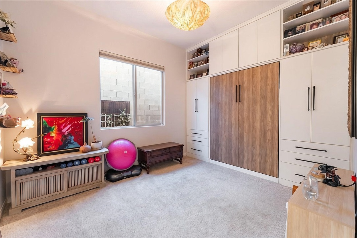 This versatile room features a large, built-in wall unit with a central wood-paneled Murphy bed flanked by white cabinetry and open shelving. The space is carpeted and includes a low console table with exercise equipment, a pink stability ball, and a wooden chest, suggesting a multi-functional use as a guest bedroom and home gym. A window provides natural light, and a unique, textured light fixture hangs from the ceiling, creating a warm and functional atmosphere.