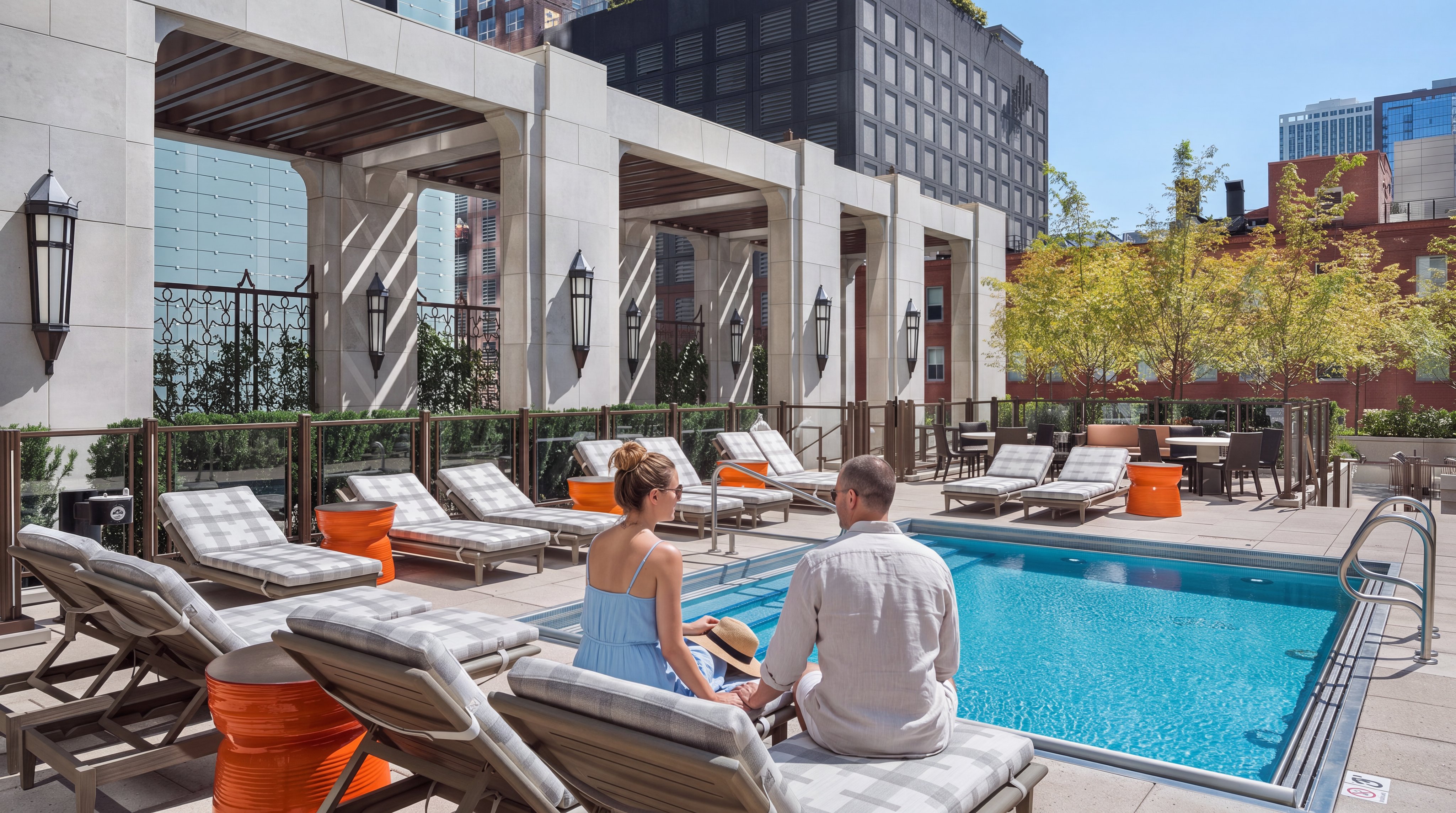 This rooftop terrace features a pristine rectangular swimming pool surrounded by numerous lounge chairs with plaid cushions. A colonnade with a pergola structure provides architectural interest and shade, while bright orange side tables add a pop of color to the space. The setting offers an urban oasis feel, complete with mature trees and a clear view of surrounding city buildings.